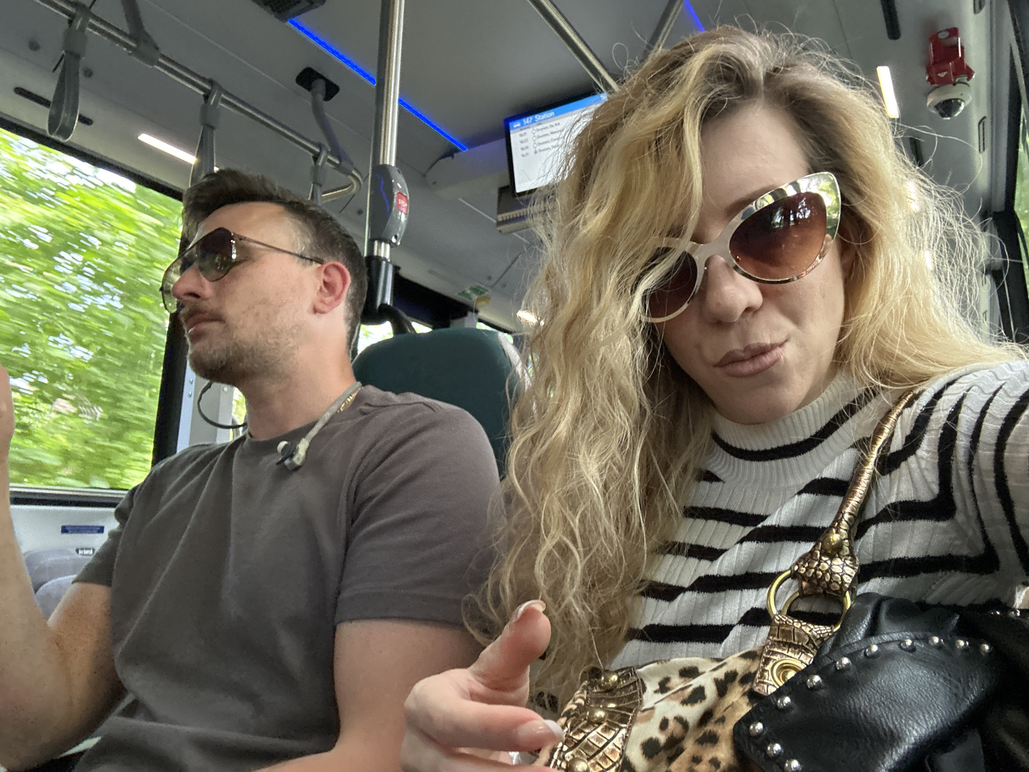 Two people are seated on a bus. A man on the left wears sunglasses and a grey t-shirt. A woman on the right wears sunglasses, a striped shirt, and has a patterned bag. The bus has a blue light strip and a display showing the route, potentially the 147 station, which includes Dronten, Netherlands. A security camera is visible on the bus ceiling. The view out the window suggests the bus is in motion.