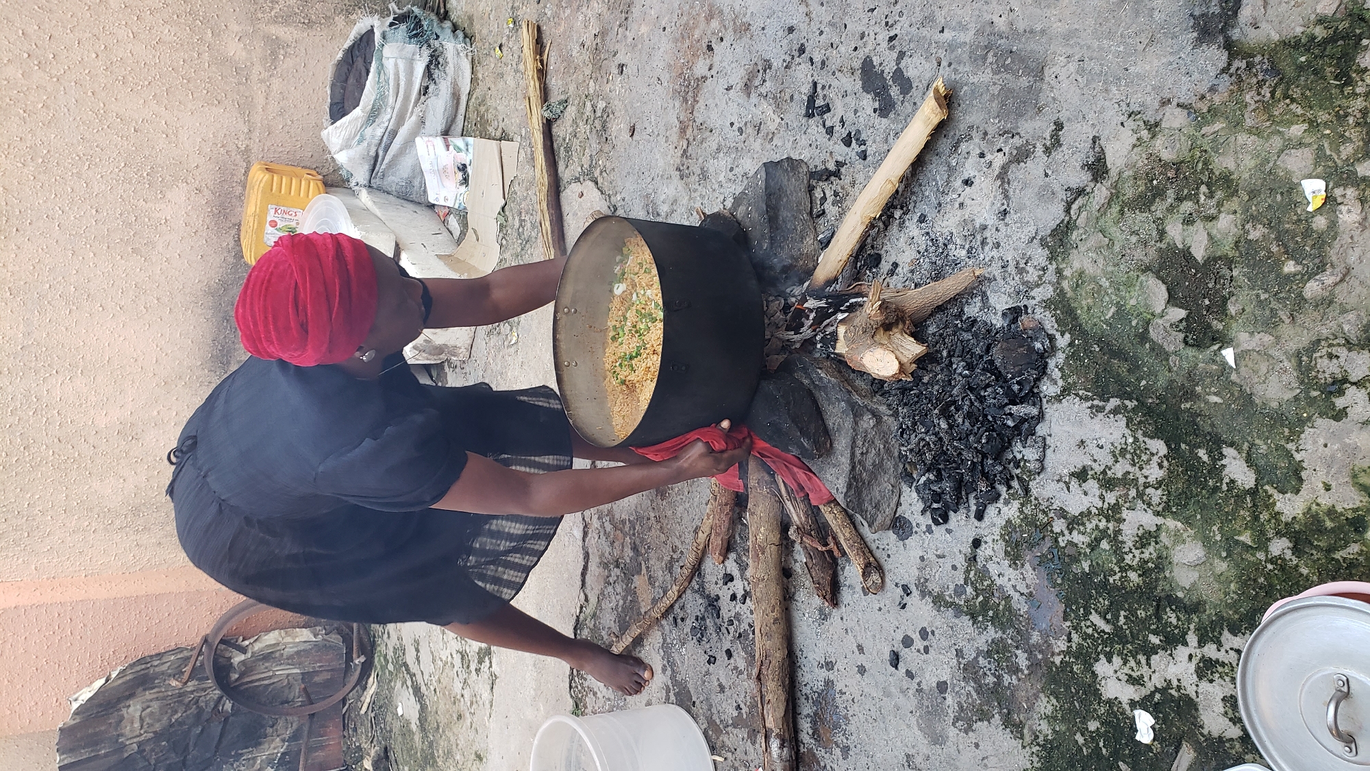 In the Sabon Tasha area of Nigeria, a woman, dressed in a black dress and a red headscarf, is cooking outdoors. She is bent over a large pot filled with rice, which sits atop a makeshift stove of stones and burning wood. The ground is made of concrete, with patches of green moss and scattered debris. In the background, various items are visible, including a yellow plastic container, a white cooler, and sacks of what appear to be goods or materials. The scene suggests a traditional cooking method being employed in an urban or semi-urban setting.