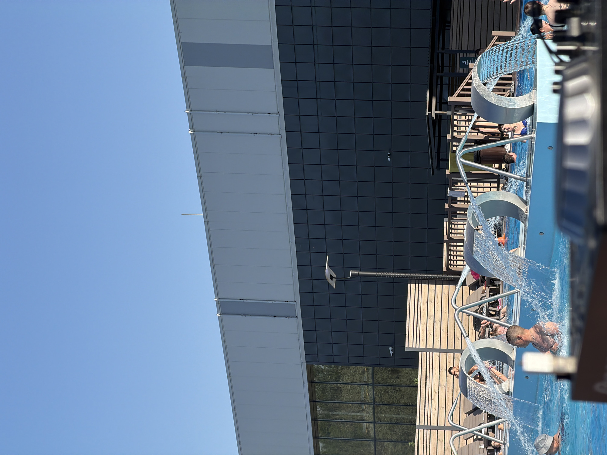 Here is a detailed description of the image:
The image shows an outdoor water park or pool area, likely in Kazan, Russia, given the user's request. The setting features a modern architectural style with a sleek building in the background, combining white and black tiled sections. Adjacent to the building is a wooden deck area equipped with lounge chairs where individuals are relaxing. The foreground is dominated by a swimming pool with several arched water features creating small waterfalls. People are seen enjoying the water, some swimming, others relaxing under the water jets. The weather appears sunny and clear, indicated by the bright blue sky visible on the left side of the image.