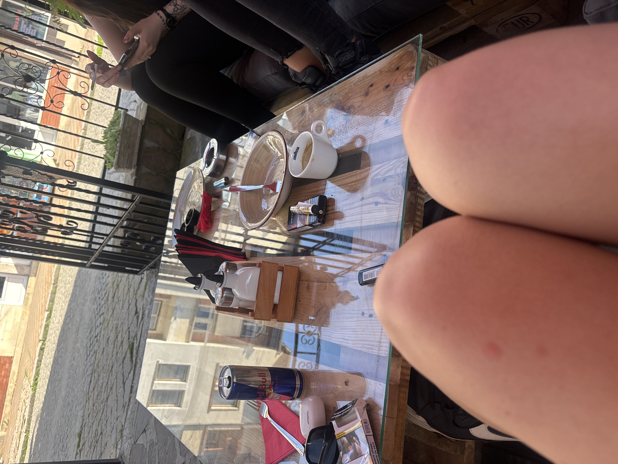 The image depicts an outdoor cafe setting in Tryavna, Bulgaria, observed from a seated individual's first-person perspective, with their knees and upper thighs visible in the lower foreground. Small red skin marks are present on the visible left knee. The person's left foot, clad in a black shoe with a white logo, is partially visible.
Seated at the same glass-topped table with a wooden base, to the right, are at least two other individuals. One person is partially visible, wearing black ripped leggings and black Adidas sneakers on their feet. Their right hand holds a lit cigarette, and their left hand operates a dark-colored electronic device, possibly a smartphone or a vaping device. A wristwatch is visible on the left wrist, and a tattoo is present on their inner forearm. Another person's legs are partially visible further to the right, also in black attire.
The glass table surface displays several items: a blue and silver Red Bull energy drink can, a red paper napkin with a silver fork, black sunglasses, a small white rectangular object, a pack of Sobranie Black cigarettes with a lighter on top, and another black rectangular lighter. A wooden caddy containing two shakers and a white bottle with a black spout (likely oil/vinegar) is positioned centrally. An empty oval-shaped ceramic bowl with a fork inside, a white Lavazza coffee cup, and a silver ashtray containing a cigarette butt are also present. The glass tabletop reflects buildings across the street, notably a light-colored building with two windows.
Beyond the table, a black decorative metal railing separates the cafe area from a cobbled stone street. Greenery grows between the pavers. A concrete planter with succulents is situated behind the railing. In the background, buildings with light-colored facades and some reddish brick elements are visible, including a white door. The scene is illuminated by direct sunlight, indicating daytime.