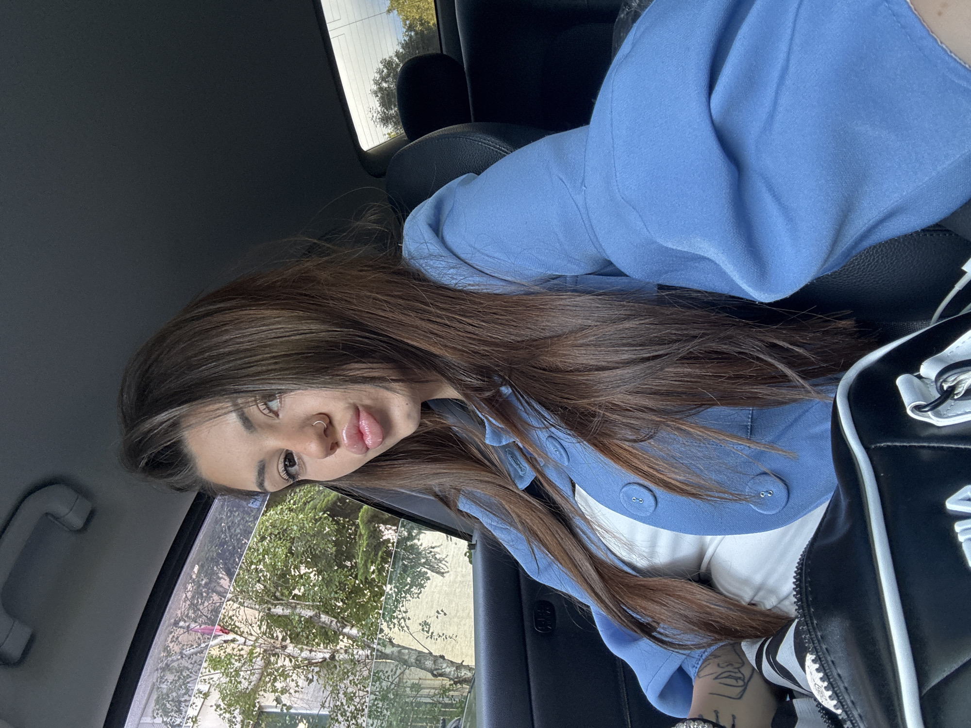 The image depicts a young woman inside a car, looking directly towards the viewer. She has long, straight dark brown hair cascading over her shoulders and chest. She is wearing a light blue long-sleeved top or jacket, featuring white fabric layered underneath and several matching blue buttons on the front. Her face is clearly visible, with full, glossy lips and a silver nose ring. A stylized tattoo of a face or profile is visible on her arm, along with a bracelet. Partially seen next to her is a black and white bag, which appears to have white lettering that could be "FILA".
The setting is a car interior, characterized by dark seating and a dark headliner. Through the car's window, verdant trees and a bright sky are visible, indicating it is daytime with clear weather. The overall mood appears casual and relaxed, with the woman posing for the camera. This scene is located in Sofia, Bulgaria.