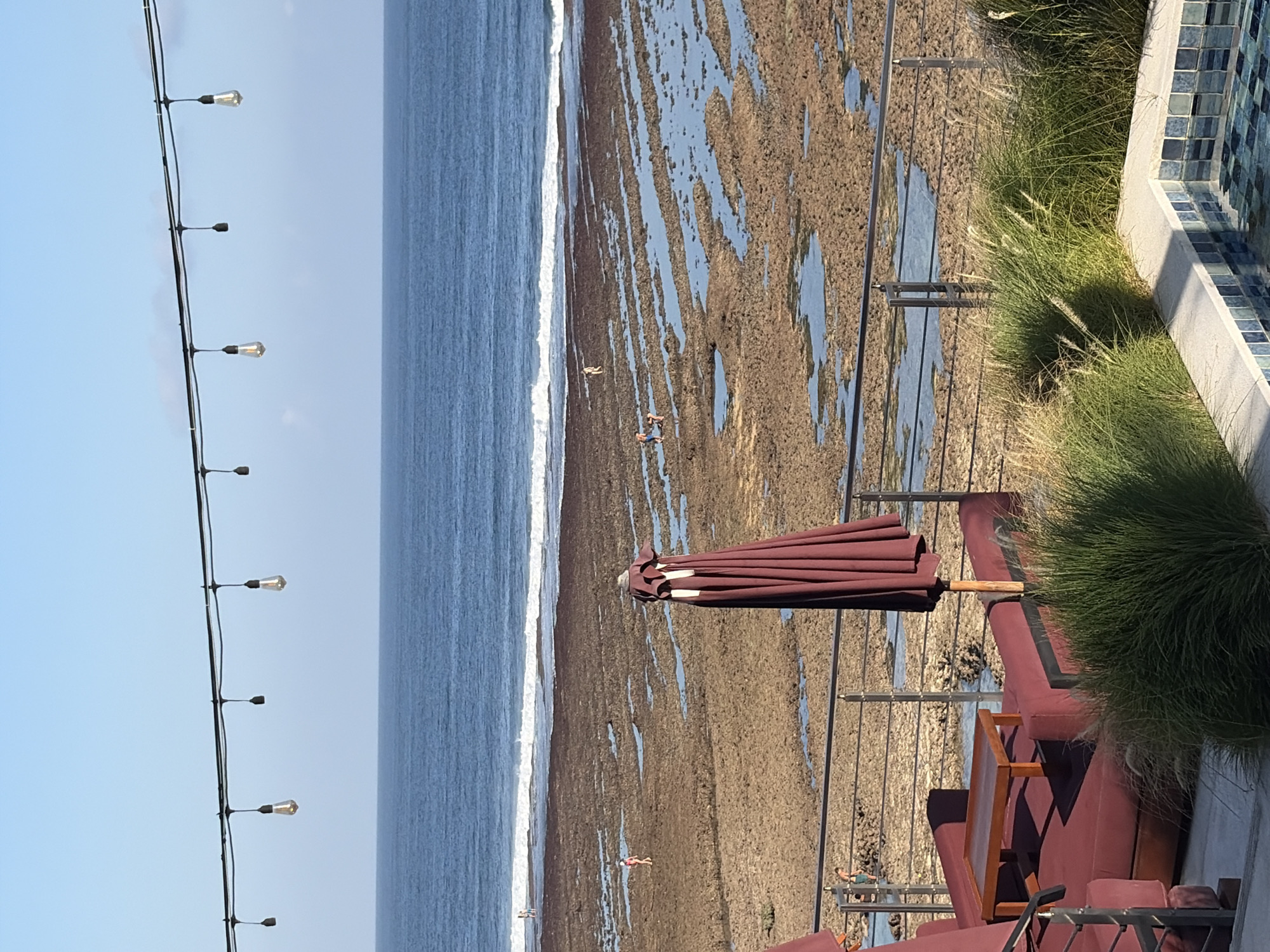 A wide shot shows the ocean from a patio in Kuta Selatan, Indonesia. The patio has maroon seating and a small wooden table. Beyond the patio is a rocky beach with tidal pools and a few people. The ocean has white waves crashing on the shore. Above the scene, string lights hang against a clear blue sky.