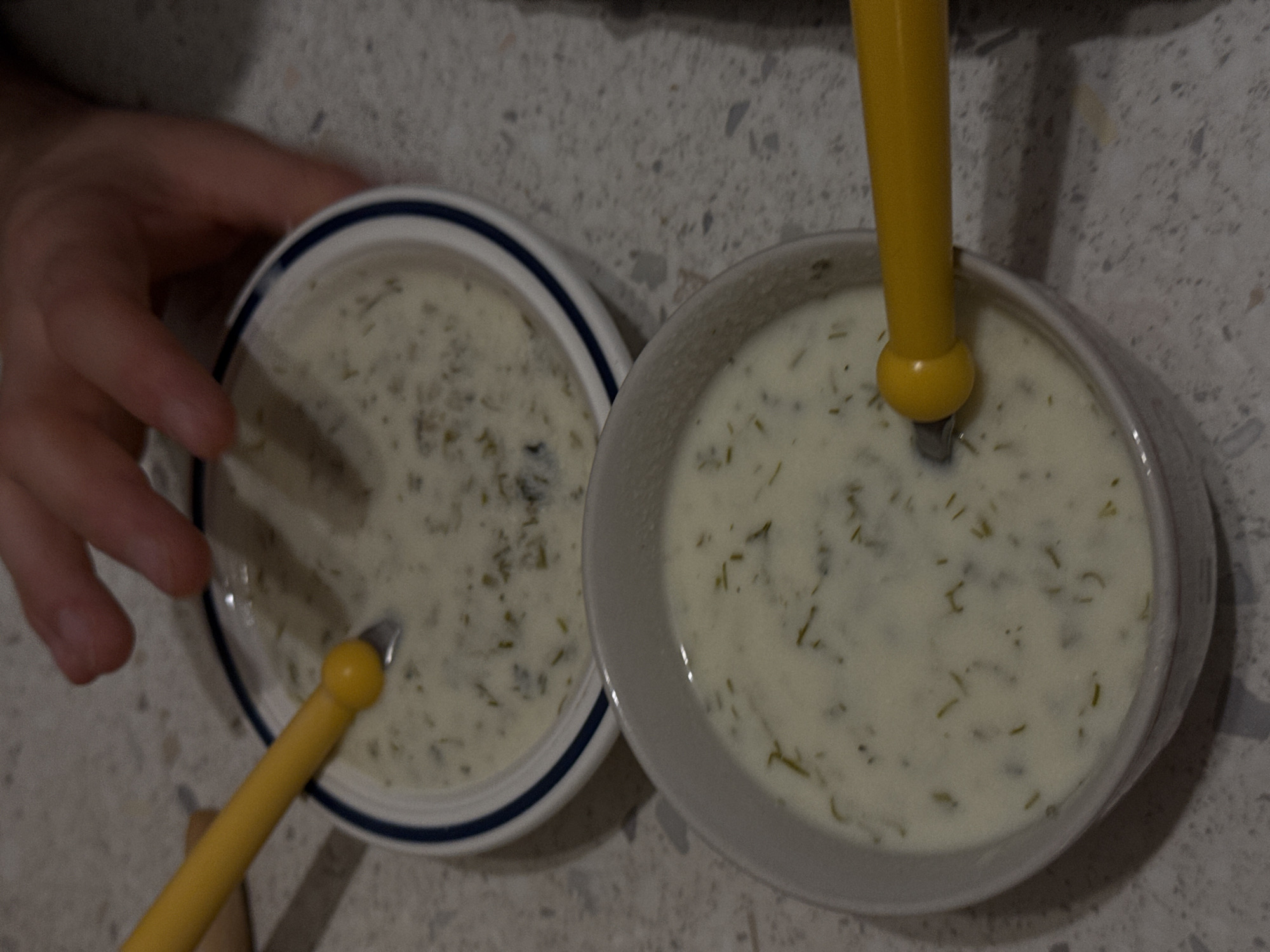 Two bowls of white soup with green herbs sit on a countertop with a speckled pattern. Yellow spoons are submerged in each bowl. A hand reaches toward the bowl on the left. No information indicates that the photo was taken in Milan, Italy.