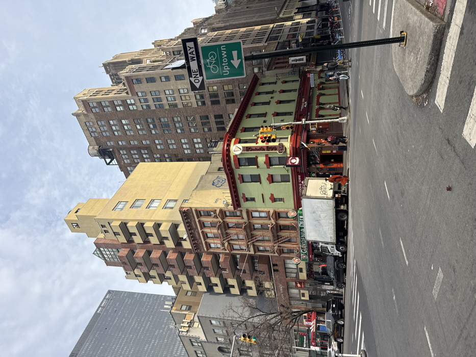 An urban street scene showcases buildings of varied heights and architectural styles. A green-painted corner building with a red trim and a bar called "Rosie O' Grady's Pub" is prominently featured. A "One Way Uptown" sign is visible. Traffic signals are placed at the intersection. Delivery trucks with the name "GARDENIA DELI" on the side are parked along the street. A taller, tan-colored building with protruding balconies stands out. Pedestrians and bikes are seen near the buildings. The street is wide with crosswalk markings.