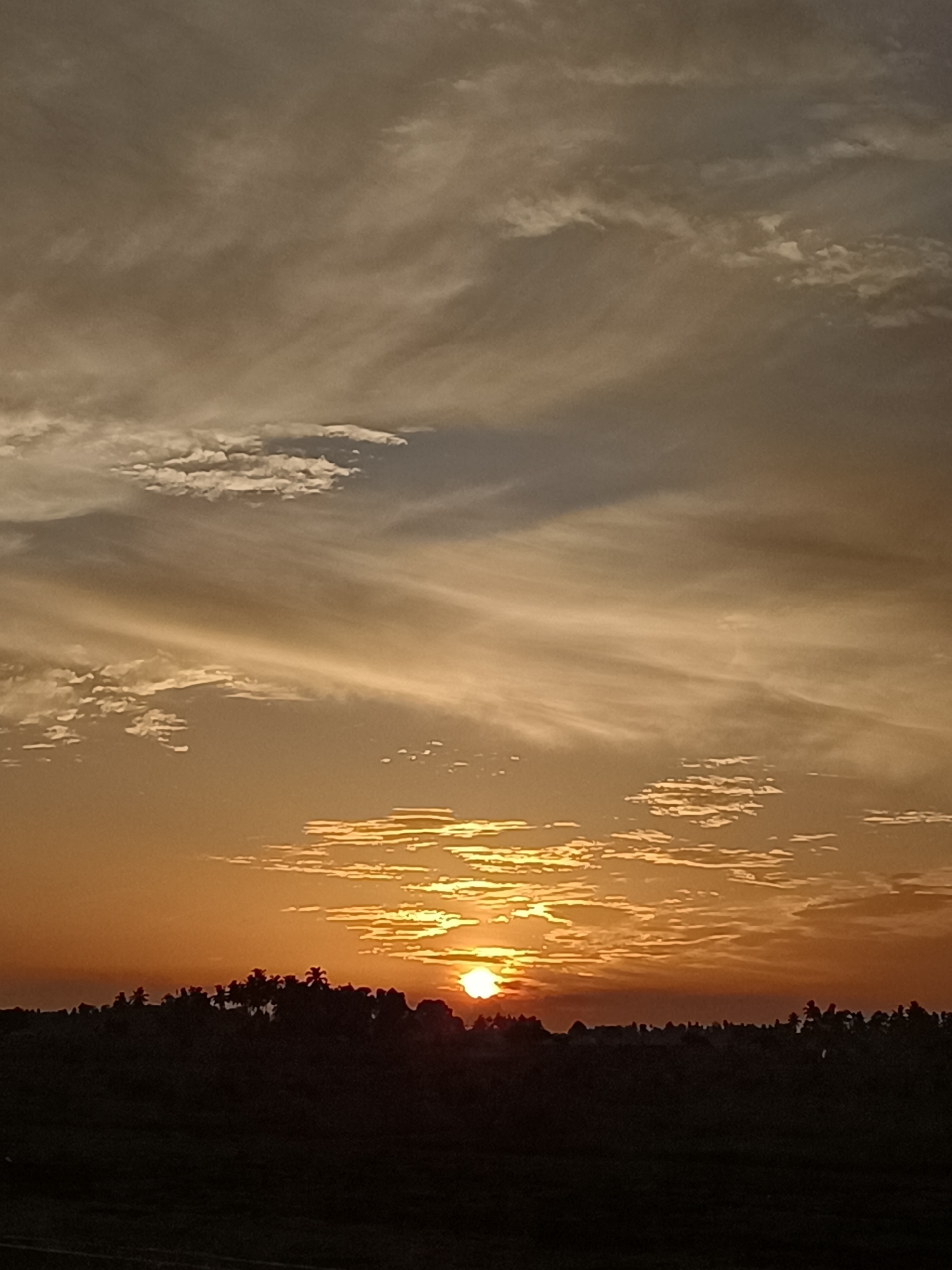 The image depicts a sunset over a silhouetted landscape in Meunasah Pupu, Indonesia. The sky is filled with various cloud formations, ranging from wispy streaks to thicker, more textured clusters. The sun, a bright orb, is partially obscured by clouds and is near the horizon, casting a warm, orange glow across the sky. Below the sky, a dark silhouette of trees and vegetation forms the foreground, suggesting a rural or natural setting. The overall atmosphere is serene and tranquil, characteristic of a sunset.