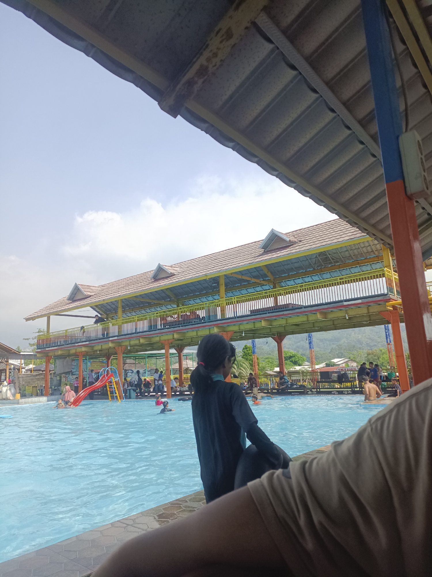 A view from under a covered seating area looking out onto a swimming pool in Prigen, Indonesia. The pool is filled with light blue water and contains several people swimming and interacting. A red and yellow slide is located on one side of the pool. A two-story building with a brown tiled roof and colorful railings overlooks the pool. Behind the building, a mountain range is visible in the distance. Several individuals are seated on the edge of the pool or walking along the perimeter. The foreground features the partial figures of people, including a girl in a dark swimsuit and a person wearing a tan shirt. The sky is partly cloudy.