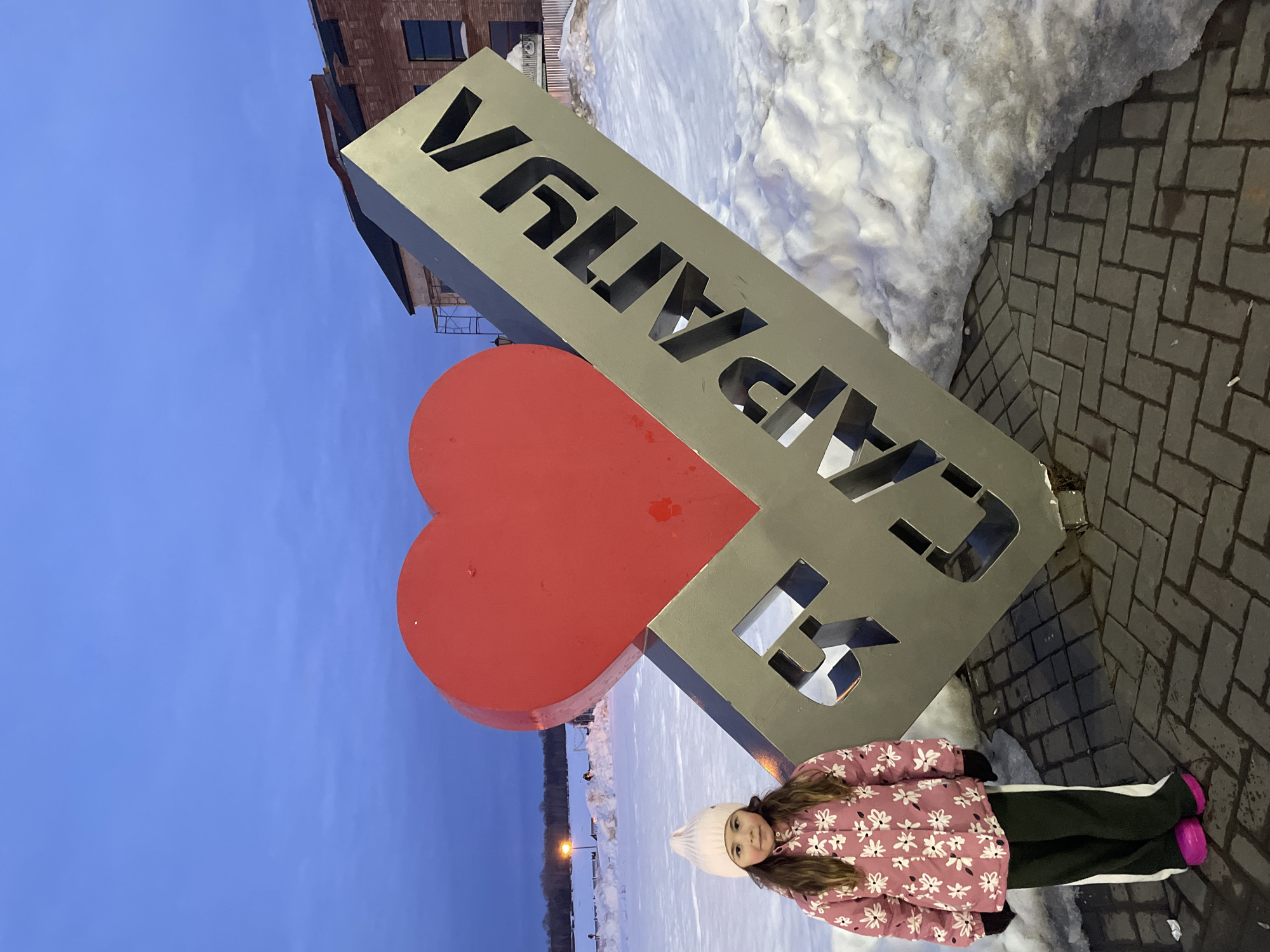 A young female child stands outdoors in Sarapul, Russia, at dusk or early evening, next to a large "I love Sarapul" monument. The monument features a prominent red heart symbol and the text "Я SARAPUL" (I Sarapul) in grey metal with cut-out black letters, positioned at an angle on a paver block base. The child, approximately 5-8 years old, has long brown hair and wears a light pink knit beanie, a pink winter jacket with a white floral pattern, dark green pants with white side stripes, black gloves, and bright pink footwear. She looks directly at the camera. The ground immediately around the monument and child consists of dark grey paver blocks, while the surrounding area is covered in substantial mounds of white snow. In the background to the left, a vast, snow-covered expanse, likely a frozen river or field, extends to a distant treeline, with a few streetlights and faint figures visible. To the right, a brick building with dark-framed windows and visible scaffolding indicates an adjacent structure. The sky is a uniform, deep blue, signifying twilight.