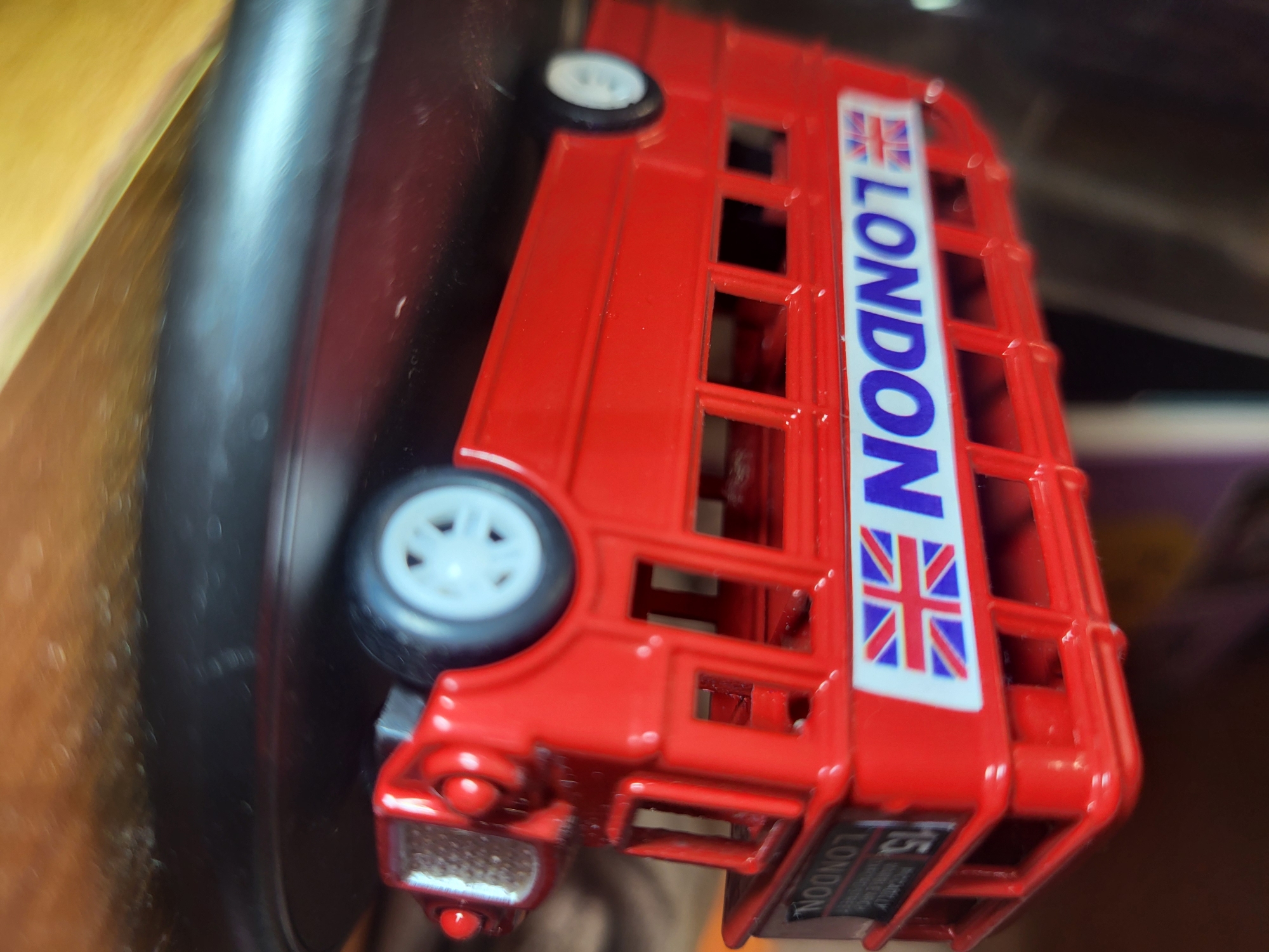 The image shows a close-up, angled shot of a red toy double-decker bus. The bus is detailed with the word "LONDON" and the Union Jack flag on its side. There is also a sign on the side of the bus that reads "15" and "LONDON". The bus is resting on a black circular base, which is placed on a wooden surface. The background is blurred, with hints of other objects and a dark, out-of-focus environment. There are no people visible in the image. The image is taken indoors.
This image does not contain any information that could be used to determine its location in the city of Horishni Plavni, Ukraine.