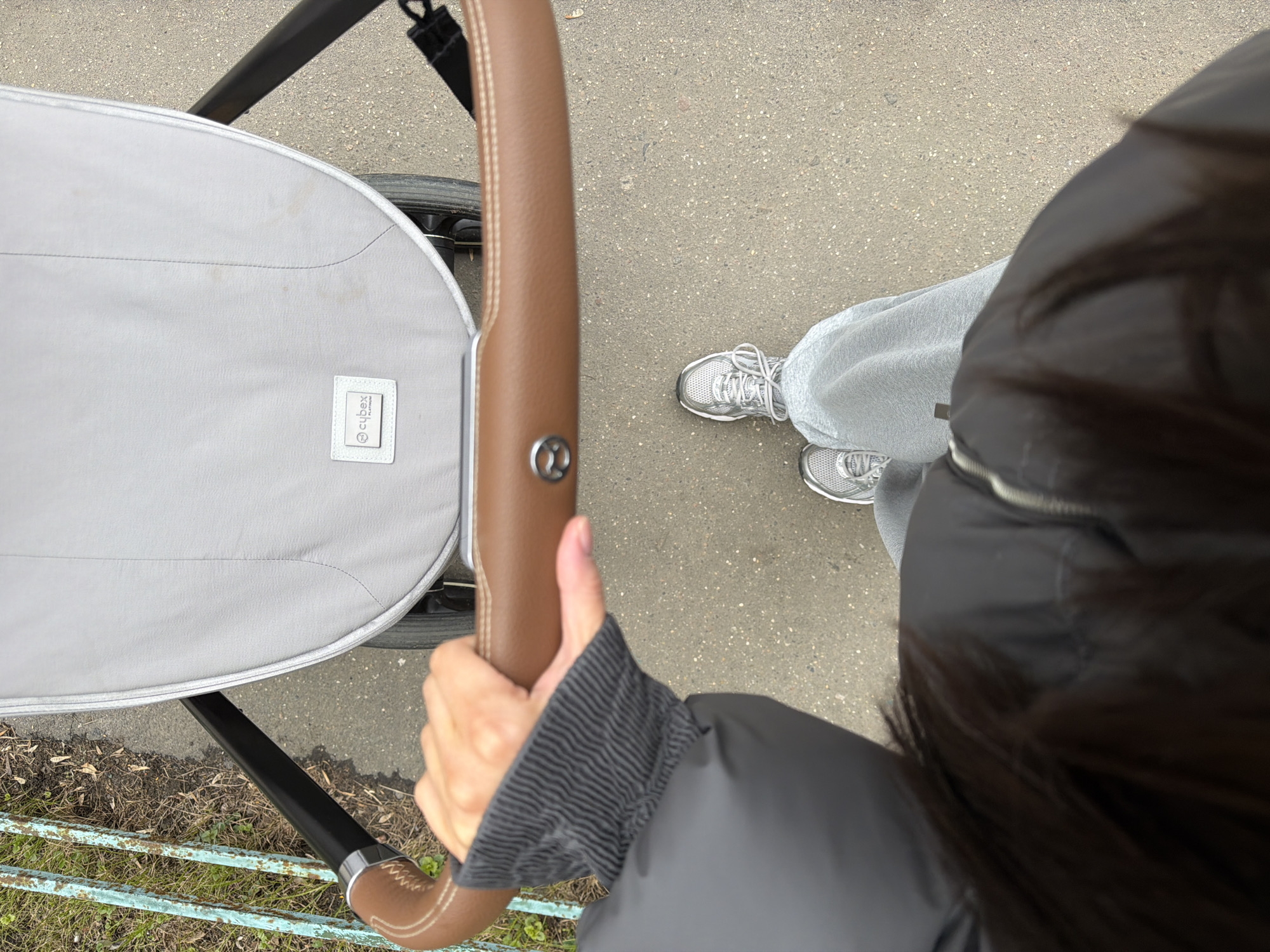 A top-down shot reveals a person holding the handle of a Cybex baby stroller on a concrete surface. The stroller is gray with a brown leather handle. The person is wearing gray sweatpants, silver sneakers, and a black jacket. Some bare earth with grass is visible in the lower left. The scene is set in an outdoor environment in what looks like the city of Khimki, Russia.