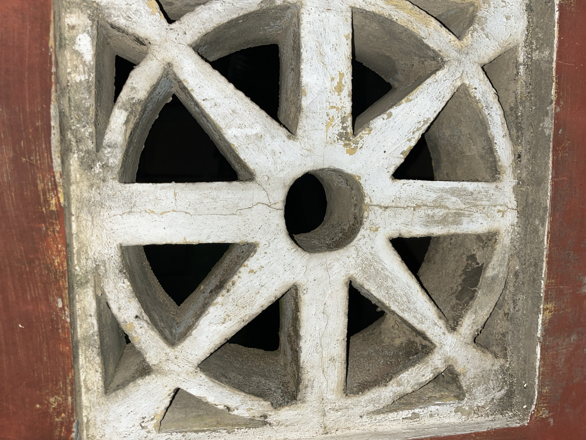 The image shows a close-up of a decorative, weathered concrete vent with a geometric pattern. The vent is white and has a circular design with spokes radiating outwards, reminiscent of a wheel or a starburst. In the center of the circle is a small, circular opening. The concrete is cracked and peeling in places, showing signs of age and exposure to the elements. The vent is set within a reddish-brown frame, likely part of a wall or structure. The background behind the vent is dark, suggesting it is a ventilation opening leading into a darker space. The lighting appears to be artificial or ambient, highlighting the texture and details of the concrete. No people or specific activities are visible. The focus is entirely on the architectural detail of the vent and its immediate surroundings. The location context suggests this might be an element found in Indonesian architecture, specifically in Kota Kuala Simpang. There is no visible text in the image.