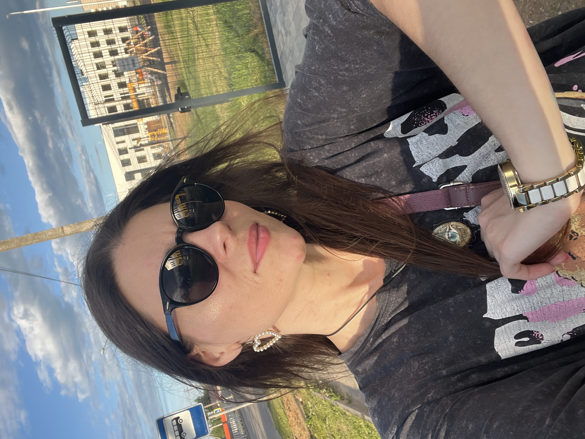A woman is taking a selfie outdoors with a cityscape in the background. She has long brown hair, is wearing sunglasses, a black t-shirt, and heart-shaped earrings. Her arm is resting on her chest, and she is wearing a watch. Behind her, a building and a playground are visible. A bus stop sign is also present. The scene appears to be in Krasnoe-na-Volge, Russia.