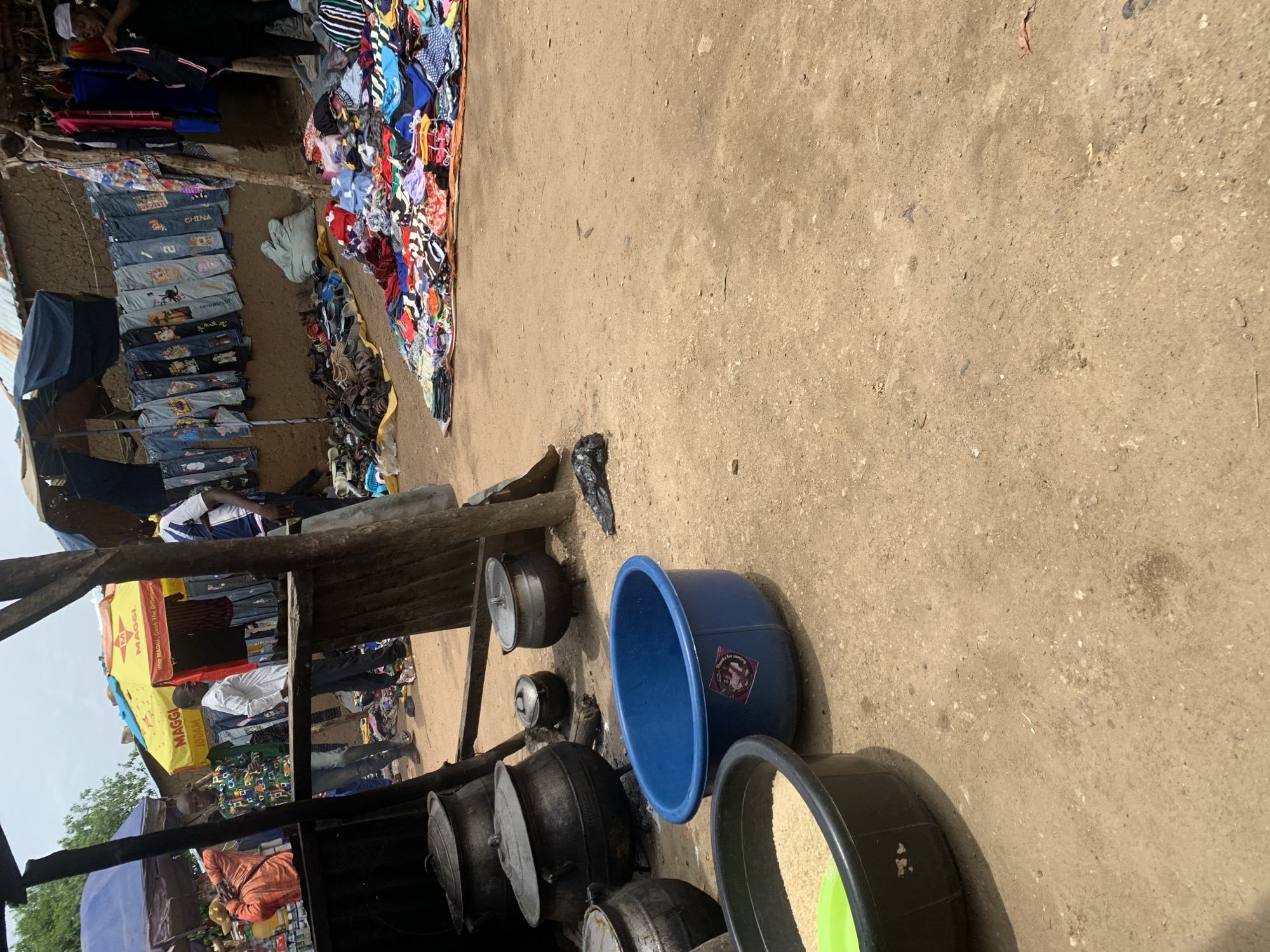 This media file captures an outdoor market scene in Nigeria, likely during daytime with clear weather conditions. The foreground is dominated by a collection of large cooking pots of various sizes, some with lids, arranged on the sandy ground. To the right of the pots, a large blue plastic basin stands prominently. Next to it, a black plastic basin is filled with what appears to be uncooked rice, with a bright green rim.
In the background, a stall is set up with a variety of clothing items displayed. Some are neatly hung on a wooden structure, while others are piled up. The clothing appears to be a mix of casual wear, including jeans and patterned tops. Several people are visible within the market. One person, wearing a white shirt, stands near the stall, while another, in an orange patterned shirt, is seated further down. A person in a blue and white striped shirt is also visible in the upper left quadrant.
A yellow umbrella with red and white branding is visible, with text that reads "MAGGI" in large letters, repeated. Below that, smaller text is visible, including "Cook The Difference". The overall impression is a bustling local market with goods for sale, including food items and clothing, and evidence of daily life activities.