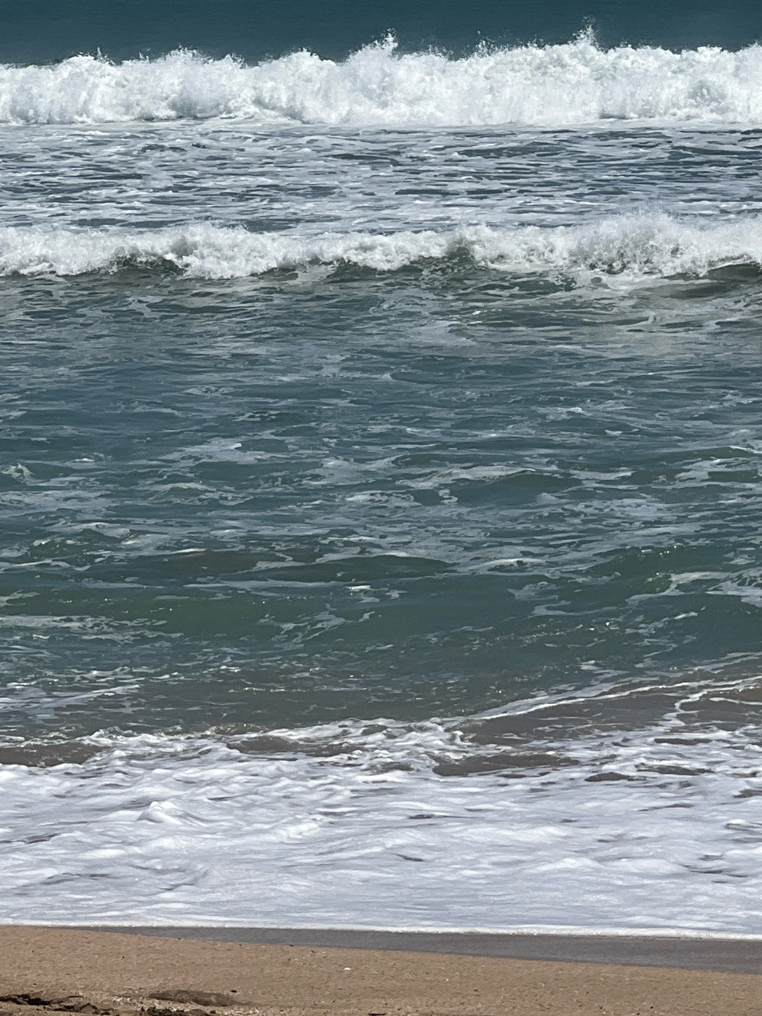 The image shows a view of the ocean and a beach. There are waves crashing on the shore, with white foam spreading across the sand. The water is a deep blue-green color. The beach is sandy and appears to be empty of people. The scene is likely a coastal area, possibly Kuta, Indonesia, known for its beaches and surf. The image is a still shot, capturing a moment in time of the ocean's movement.