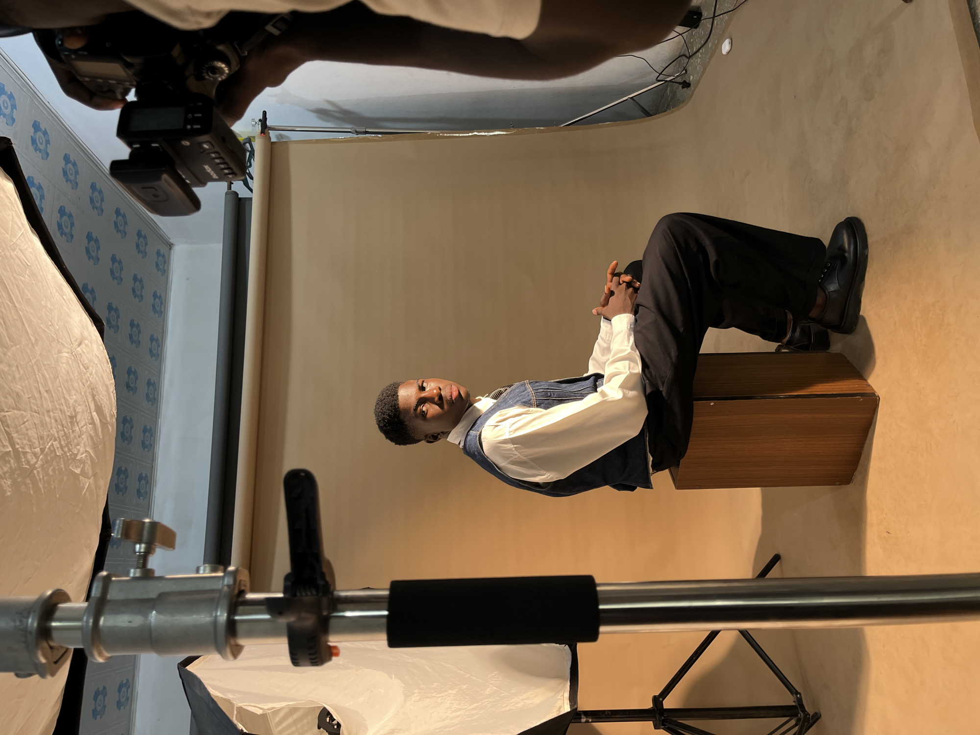 The image shows a behind-the-scenes view of a photo shoot. A young man is seated on a wooden box in front of a beige backdrop, wearing a white shirt, a denim vest, and dark trousers with black shoes. His hands are clasped together, and he is looking towards the camera. A photographer's hand is visible at the top of the frame, holding a camera. A softbox is positioned to the left of the frame, supported by a metal stand. The background consists of a wall with a light blue and white patterned wallpaper on the left and the beige backdrop on the right. The location is Unknown, Unknown.