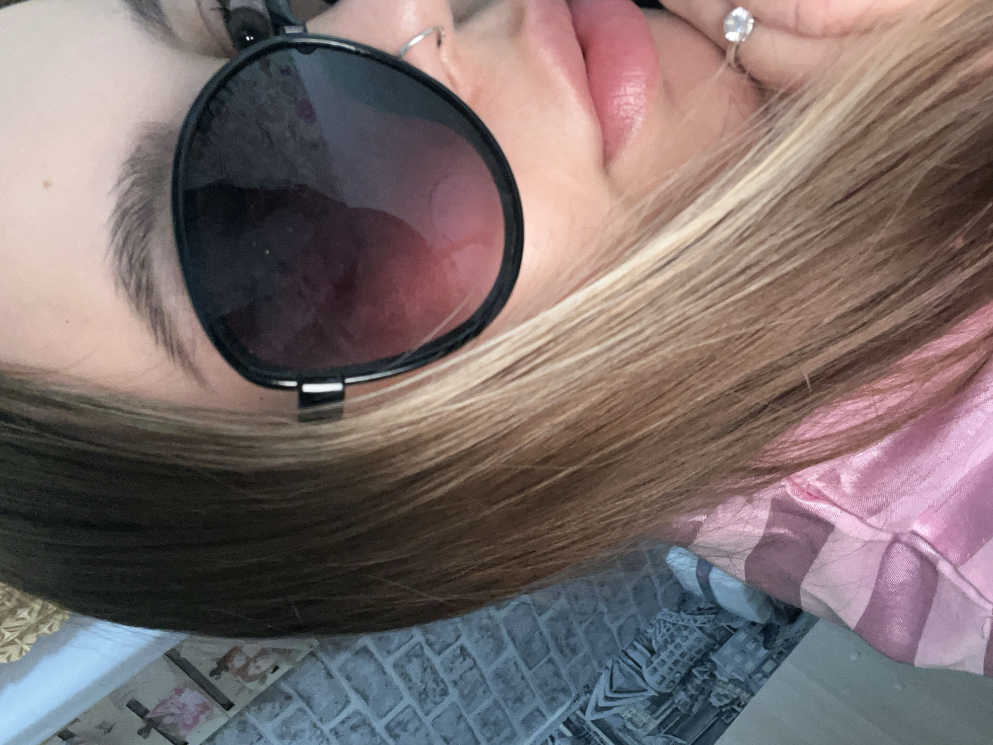 A close-up photograph depicts a person's face partially obscured by sunglasses. The person has fair skin, full lips with light pink lipstick, and blonde hair with brown undertones. A nose ring is visible in the left nostril and a ring with a prominent stone adorns the ring finger of the hand. The person is wearing a pink and white striped garment. The background is blurry but shows what appears to be a surface. The photo lacks context to determine if this picture was taken in Novopokrovskaya, Russia.