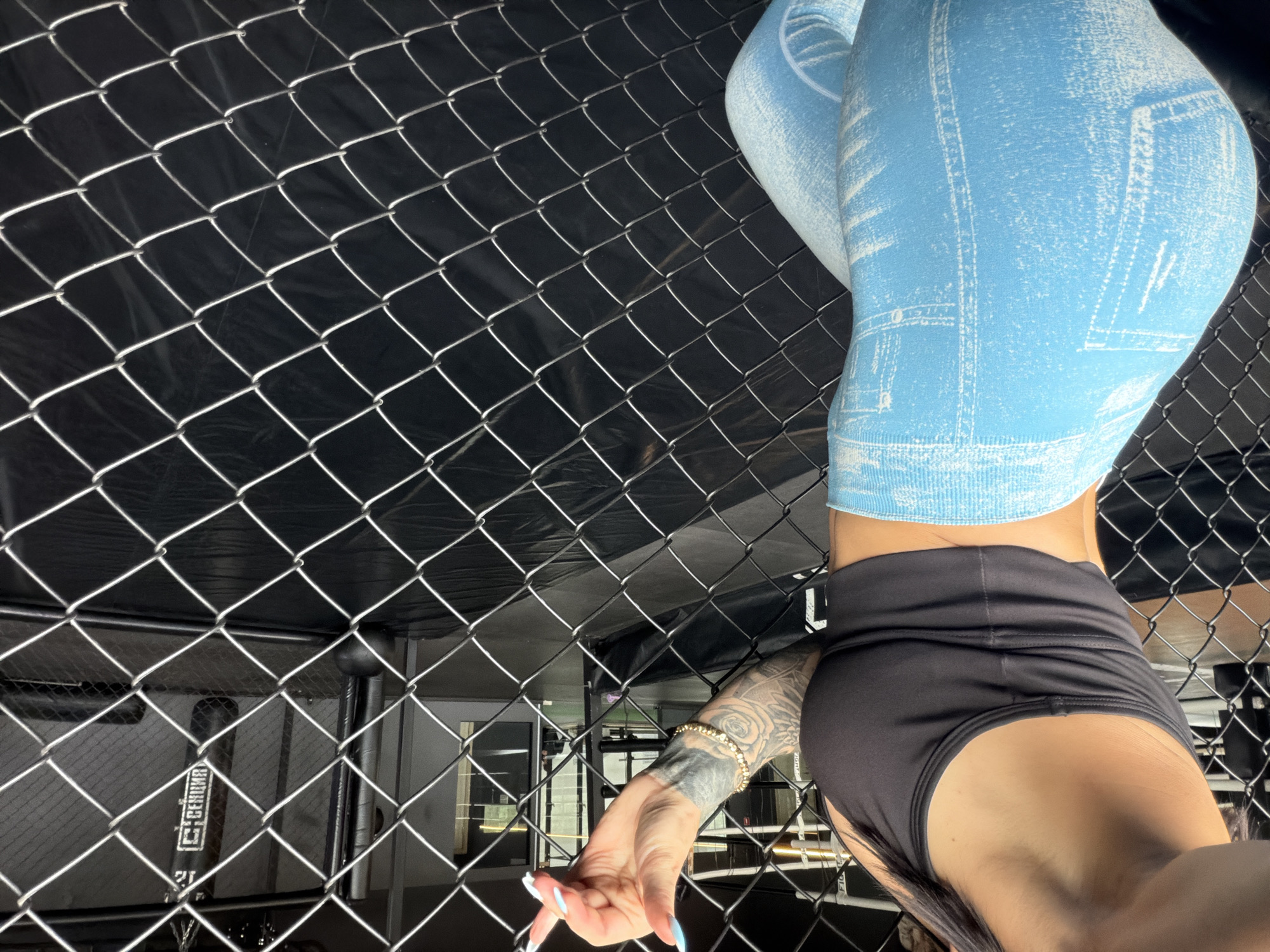 The image depicts a person performing a handstand in a fighting cage. The person is wearing blue leggings with a denim print and black shorts. The background is the interior of a building, likely a gym or arena, with a chain-link fence of the cage prominently in the foreground. The lighting suggests an indoor setting, possibly at night or in a dimly lit space. The person's body is oriented upside down, with their legs towards the top of the frame and their head and torso towards the bottom. Visible tattoos are on the person's arm, and they are wearing a bracelet. The person's hands are reaching out, one with painted fingernails. A baseball bat with visible text "GERMAN" is leaning against a structure behind the fence. The scene appears to be in Ufa, Russia.