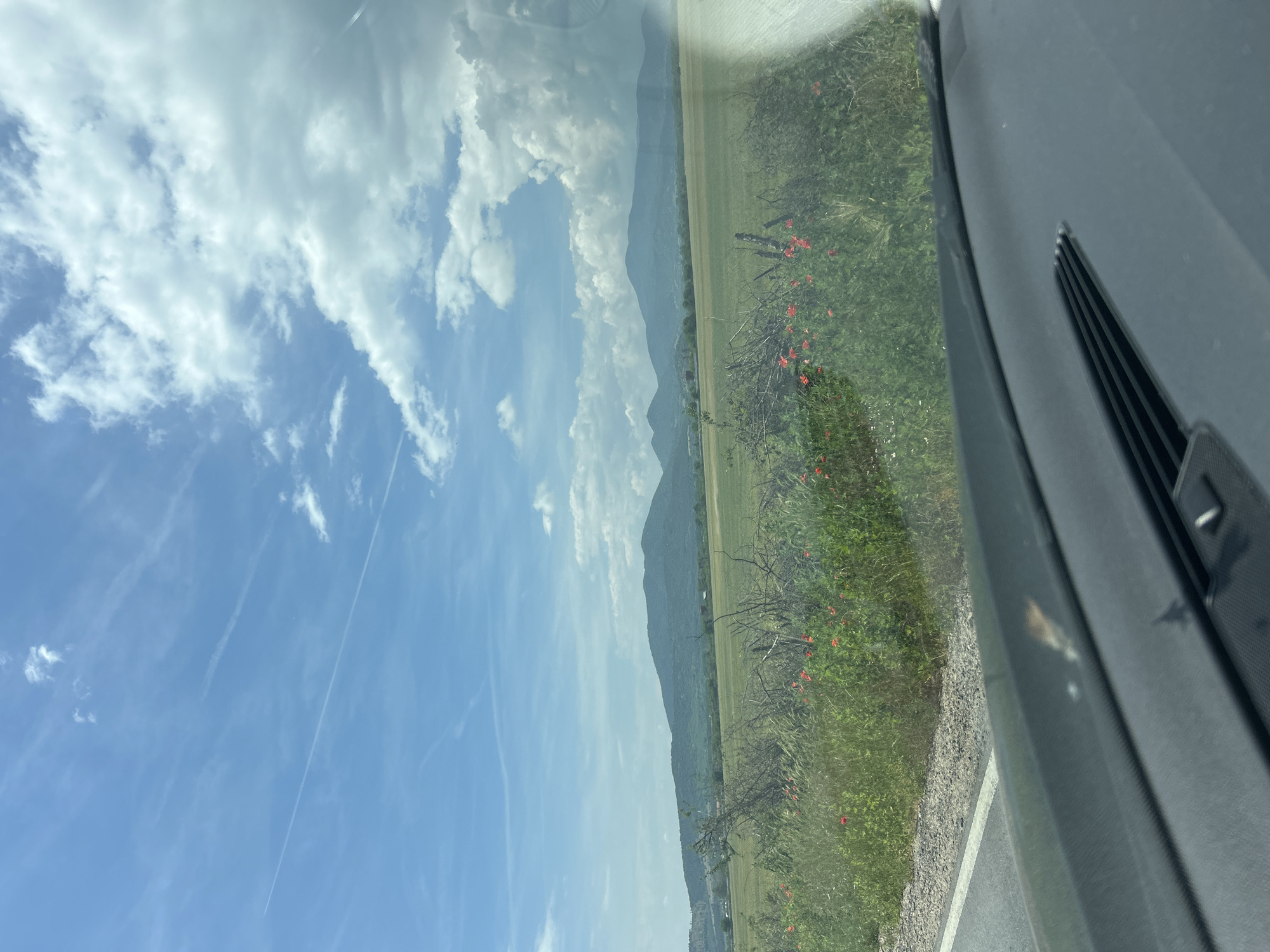 This image captures a vibrant rural landscape viewed from inside a moving vehicle, likely a car. The scene is dominated by a clear blue sky interspersed with fluffy white cumulus clouds and faint, wispy contrails from aircraft.
Below the sky, a long range of mountains stretches across the mid-ground, appearing hazy in the distance under the bright daylight. The land in front of the mountains consists of vast, flat green fields, suggesting agricultural use.
In the foreground, a strip of roadside vegetation is visible, featuring lush green grass and bushes. Bright red flowers, possibly poppies, are scattered throughout this greenery, adding a pop of color. A paved road with a distinct white edge line runs along the very bottom right. The immediate foreground on the right side of the image shows parts of a car's interior, including a dark-colored dashboard and an air vent with horizontal slats, indicating the photograph was taken through a side window. Reflections on the window also partially obscure the view.
The time of day appears to be midday or early afternoon, characterized by strong sunlight and clear visibility. The weather is fair with scattered clouds. The image provides a glimpse of a scenic journey through a mountainous, cultivated region, consistent with the provided location context of Marino Pole, Bulgaria. There is no visible text in the image.