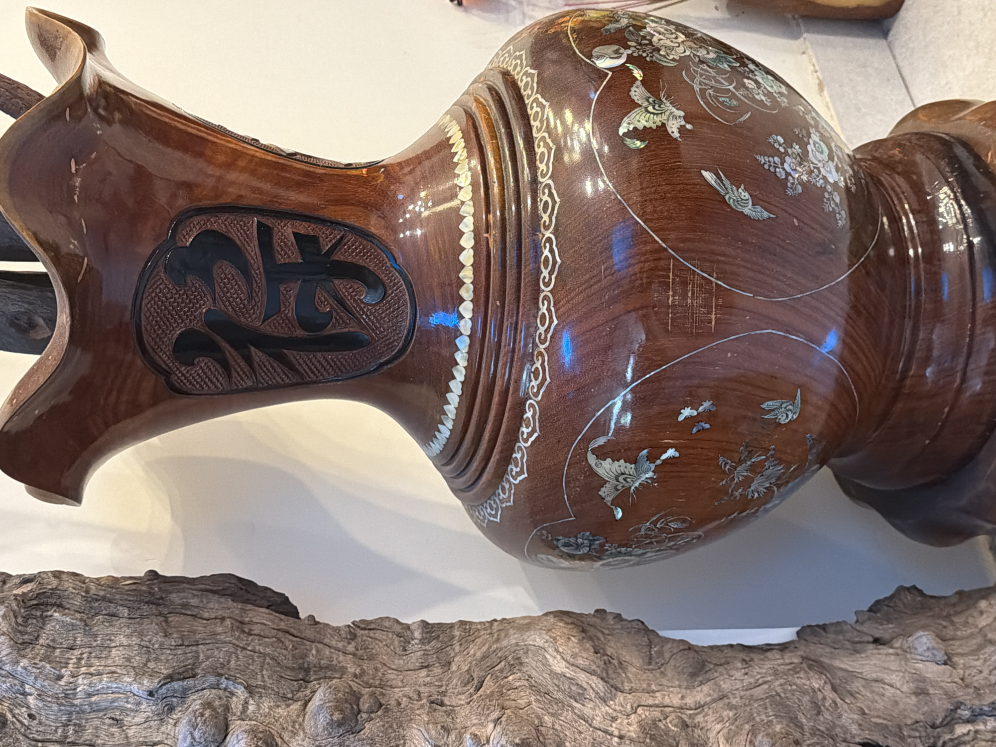 A polished wooden vase, likely part of an ornate decorative piece, is displayed. The vase is a rich brown color and features intricate mother-of-pearl inlay work depicting birds and floral motifs. A band of inlaid geometric patterns runs around a section of the vase, and a carved dark symbol, possibly a Chinese character, is prominently featured on a recessed oval panel. The vase is positioned horizontally, resting on a piece of gnarled, weathered wood. The background is a plain white wall, suggesting an indoor display. The lighting appears to be artificial, casting soft reflections on the glossy surface of the vase. The scene suggests a close-up view of a handcrafted item, emphasizing its detail and artistry. The vase itself is curved and bulbous in shape, with a narrow neck and base. The overall impression is one of elegance and traditional craftsmanship. No people are visible in the image. The time of day and weather are not discernible. The location context is Bac Nha Trang Ward, Vietnam.