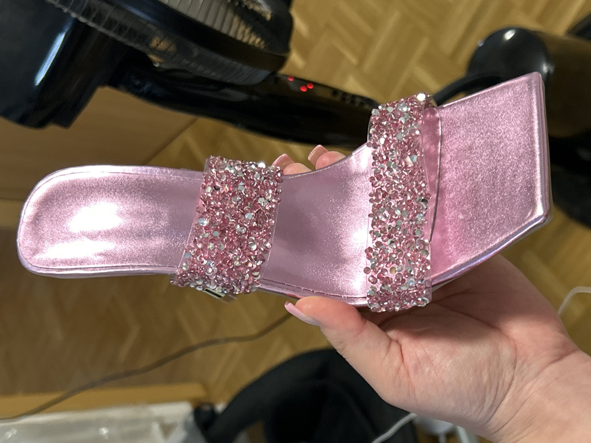 An individual holds a single light pink, square-toed high-heeled mule sandal in an indoor setting in Waldbröl, Germany. The sandal features a metallic finish and two translucent straps across the vamp, densely embellished with pink and clear rhinestones. A light-skinned hand, exhibiting an elongated, pointed, light pink manicured thumb nail, grasps the inner arch of the sandal. The background consists of a light brown wooden floor with visible plank patterns. On the far right, a large, dark-colored standing appliance, possibly an oscillating fan, is visible, displaying a black grille and three illuminated red indicator lights on its body. A light-colored vertical surface and dark electrical cables are discernible on the far left.