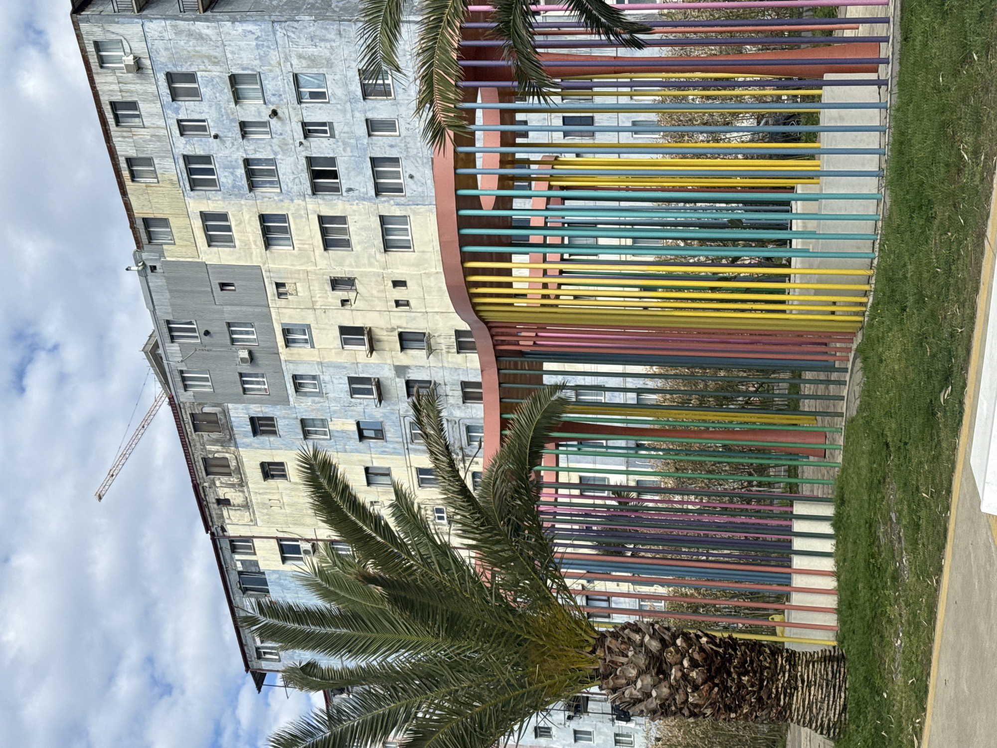 The image captures a vibrant urban scene in Batumi, Georgia, featuring a tall, somewhat aged apartment building, a colorful artistic fence, and a prominent palm tree.
On the left side, a multi-story building rises, its facade a mix of faded blue, gray, and yellow sections, some appearing to be corrugated metal siding. Numerous rectangular windows, some open, some closed, are visible, along with external air conditioning units. A faint construction crane arm is discernible against the partially cloudy blue sky in the upper left background.
Running horizontally across the mid-ground is a distinctive artistic barrier composed of numerous vertical cylindrical poles. These poles are painted in a striking spectrum of bright colors including red, pink, purple, blue, teal, green, yellow, and orange. The top edge of this colorful structure exhibits a gentle, undulating curve.
A large palm tree stands in the foreground, its robust, textured brown trunk at the bottom and its lush green fronds reaching upwards and extending over the colorful barrier. To the right of the barrier, a strip of vibrant green grass borders a concrete sidewalk, which features a distinct yellow painted line along its edge.
The scene is set during the daytime under a partially cloudy sky, showcasing a blend of functional architecture and decorative public art. No people are visible, and there is no discernible text.
