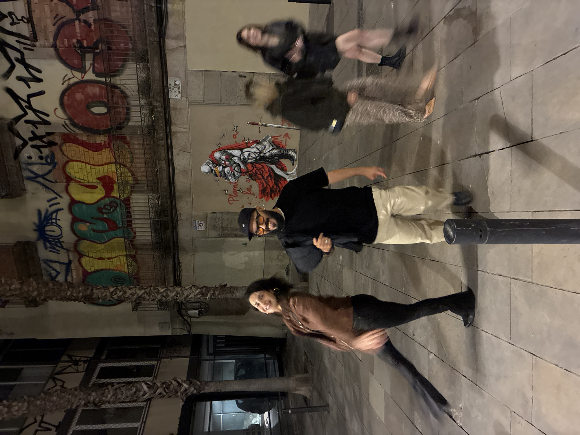 The image depicts a nighttime street scene in Barcelona, Spain. Four people are present, blurred indicating movement. A man stands in the center with a black shirt and light-colored pants. He wears a black cap and appears to be looking at the camera. A woman in a brown jacket and black pants is in the foreground. Two more blurred individuals are in the background.
Graffiti covers the wall of a building, including stylized text and an illustration of a knight. A street sign is visible above the knight. The ground is tiled. A cylindrical object or post stands near the woman in the foreground. Foliage of a tree is located to the left. A building with windows is partially visible in the background.