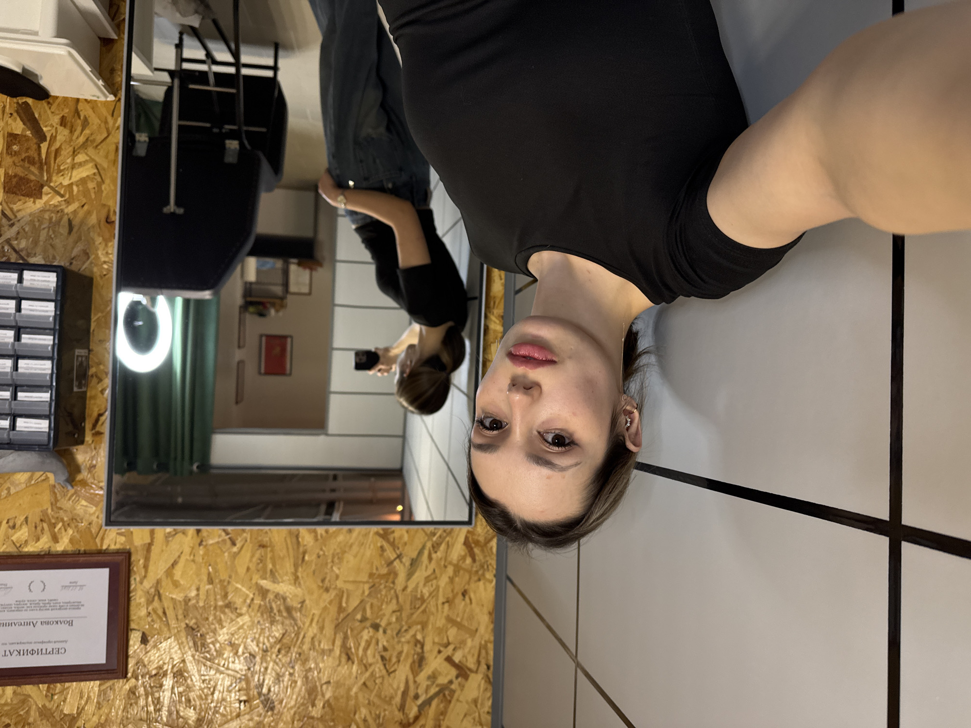 A selfie taken by a fair-skinned woman wearing a black shirt and a small earring is shown. The photo is rotated, but the woman is facing forward. In the background, a mirror reflects another person, also wearing a black shirt and jeans, holding a phone. The mirror is positioned against a wall constructed from oriented strand board, with a certificate framed and mounted on the wall beneath the mirror. Other items visible in the mirror's reflection include a ring light, green curtains, a dark chair, and various objects on shelves. The selfie taker is positioned against a white wall with black trim. The city location is unknown.