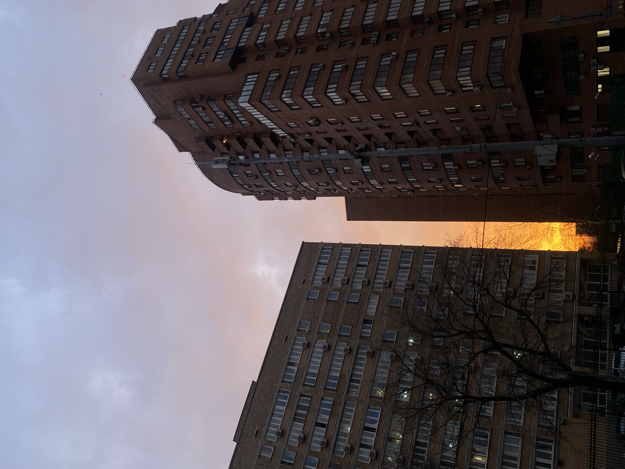 An exterior shot depicts two multi-story residential buildings in Moscow, Russia, at what appears to be either sunrise or sunset. The sky above is a gradient of lavender, pink, and orange hues. To the left, a rectangular building with a light beige brick facade features numerous windows, many of which have individual external air conditioning units mounted below them. Several windows show illuminated interiors. In front of this building, a bare-branched deciduous tree is silhouetted against the ambient light. To the right, a taller, dark red brick building with a prominent curved facade and numerous windows also displays multiple external air conditioning units. Cell tower equipment is affixed to a vertical pole on its left side. A third, darker building is partially visible behind it. A black metal fence runs across the foreground, with partially obscured vehicle shapes visible behind it. A streetlight pole with attached electrical boxes stands between the two main buildings. No people, discernible actions, or interactions are visible.