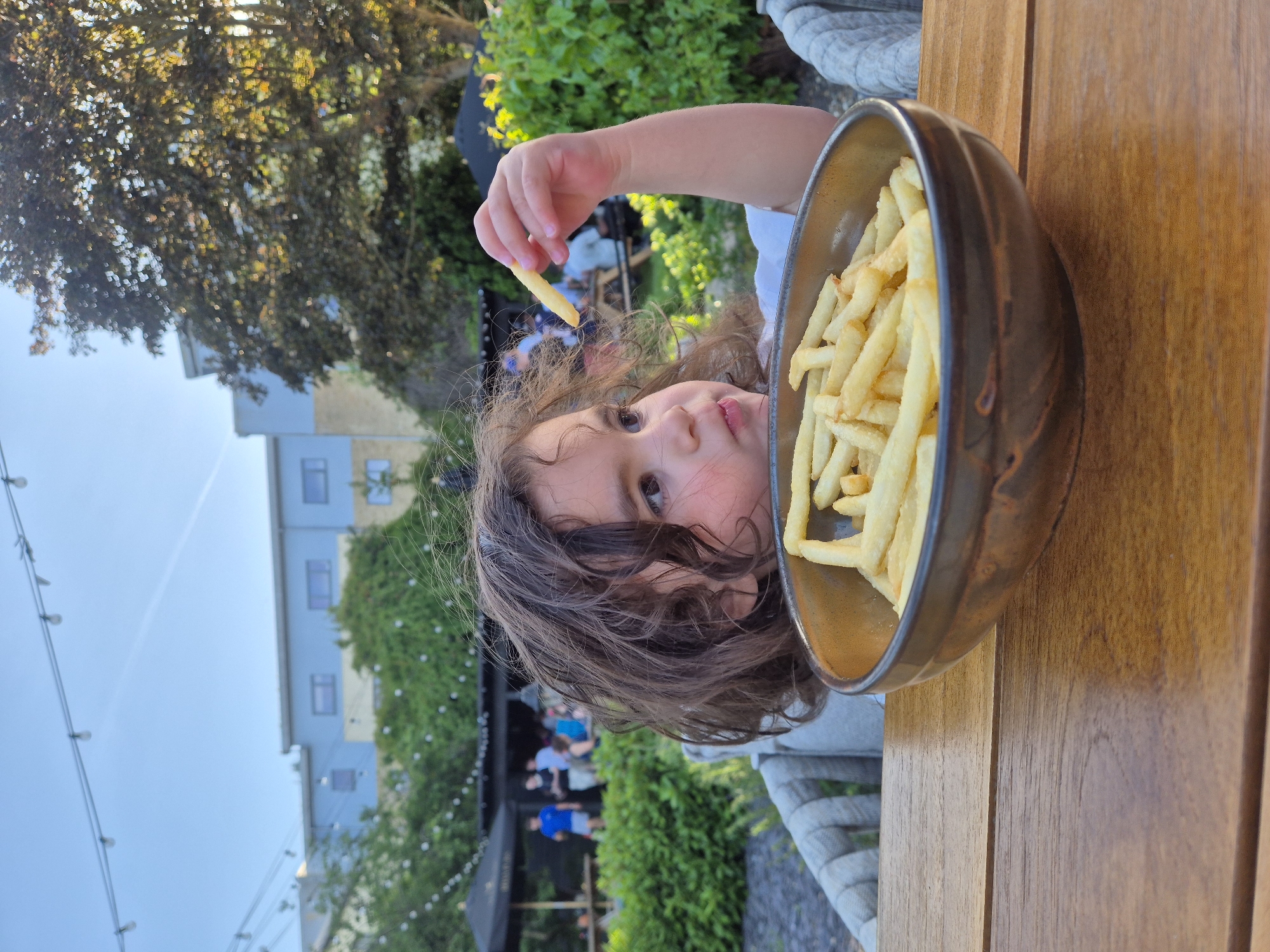 A child with brown curly hair is holding a single french fry above a bowl filled with french fries on a wooden table. The child's face is visible, and they appear to be looking at the french fry. The background includes a blurred view of a building with greenery, trees, and other people in the distance. The location is West Suffolk, United Kingdom, based on the implied geographical location.