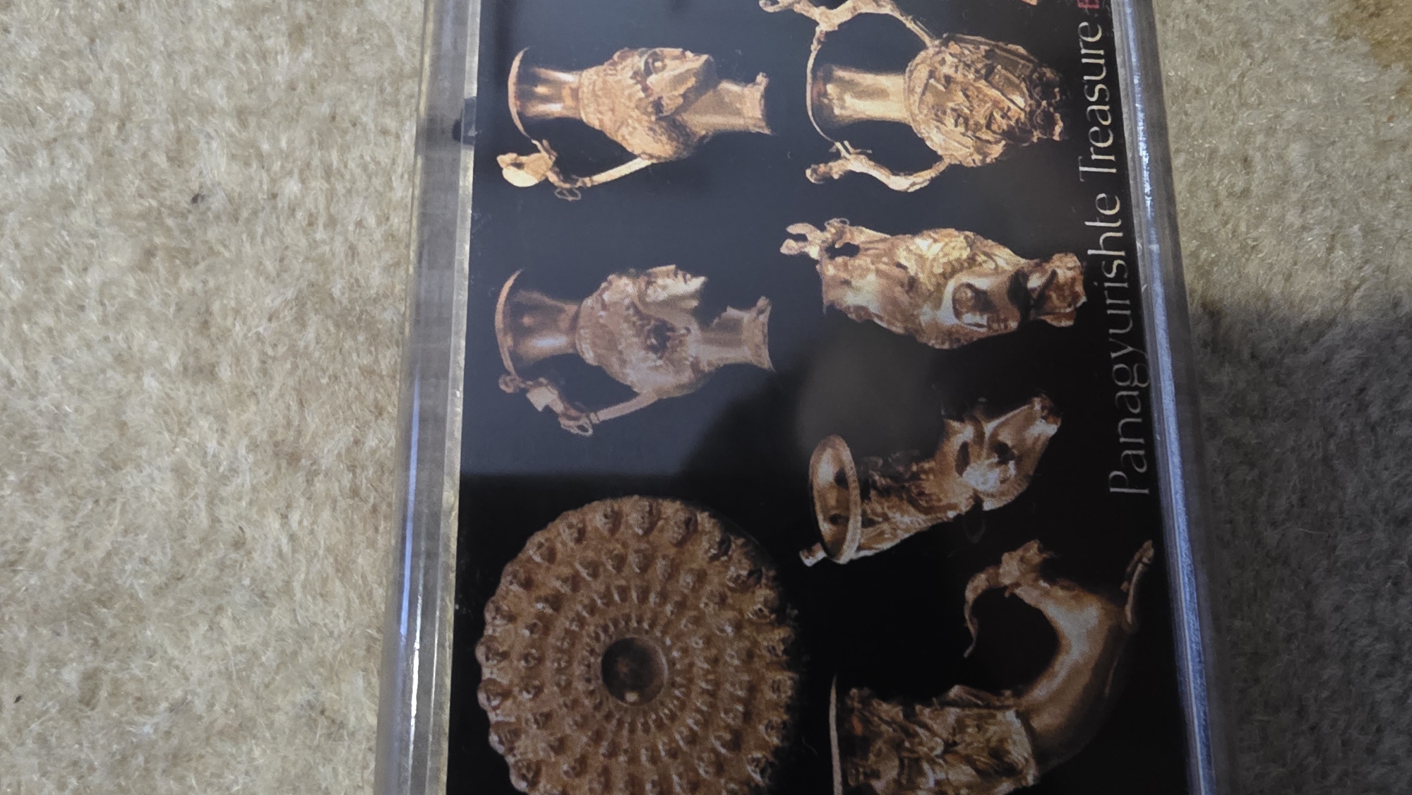 The image shows a framed picture of the Panagyurishte Treasure, a collection of Thracian golden artifacts. The picture is primarily black, displaying various golden vessels and a flat, intricately designed plate. The frame appears to be clear or silver-colored. The background visible to the side of the framed picture is a fuzzy, light-colored surface, likely a carpet or rug. There are no people visible in the image. The objects in the framed picture are the focus. The city of Blagoevgrad, Bulgaria is not depicted in this image.