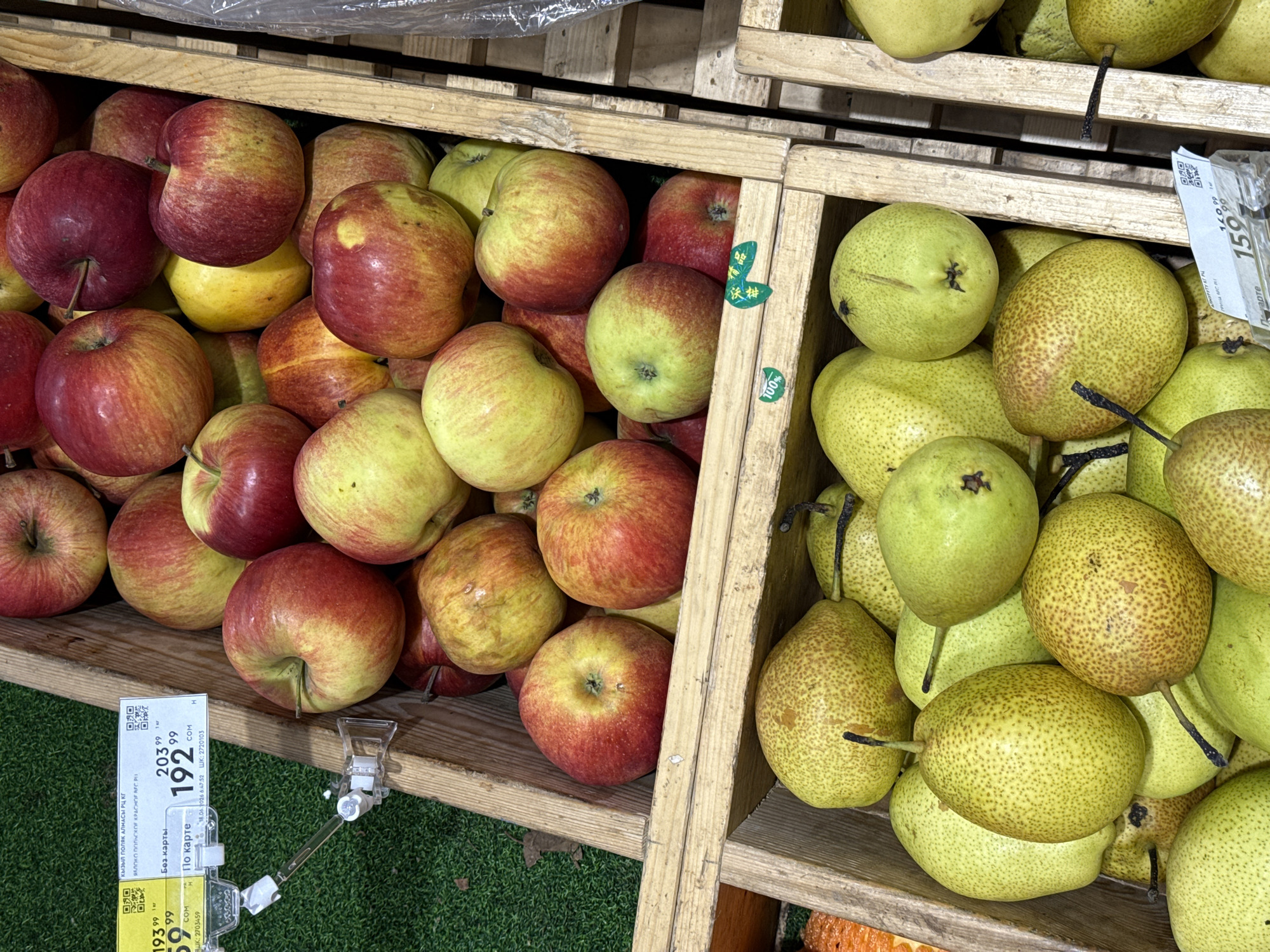 A close-up, eye-level shot shows two wooden crates overflowing with fresh fruit, likely in a market or grocery store. The upper crate is filled with red and yellow apples, some with streaks of red and yellow, while others are predominantly red or yellow. The lower crate contains a bounty of green pears with speckled brown skin and dark stems. The background on the left is a blurred green surface, possibly artificial turf or a green mat. A price tag is visible attached to the upper left of the frame, displaying numbers and text in Cyrillic. Another price tag with Cyrillic text and a barcode is partially visible in the lower right of the frame. The lighting is even, highlighting the textures and colors of the fruit.