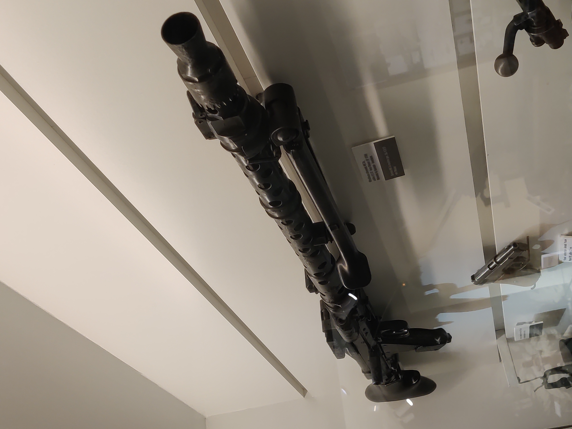 In Dunkirk, France, a machine gun is displayed in a museum. The machine gun is mounted on a wall and appears to be an MG 42, a German WWII-era weapon. It is dark in color and has a long barrel with cooling vents. A smaller handgun is also visible below the machine gun, also displayed on the wall. The background consists of white walls with angled lighting.