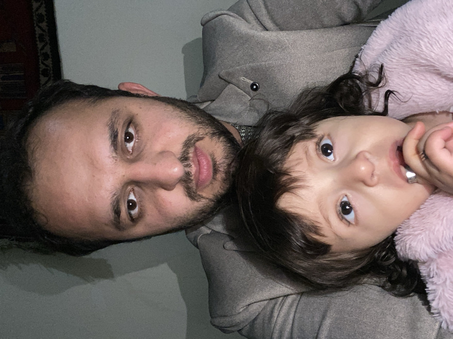 A man and a young child lie together, closely positioned, appearing to pose for a photograph. The man has dark hair and a beard, wearing a grey overcoat. The child has dark hair, big, expressive eyes, and wears a pink fluffy garment. The angle is slightly tilted, with the man's face appearing on the left side of the frame and the child's face on the right. The child's hand is near their mouth. The background is plain.