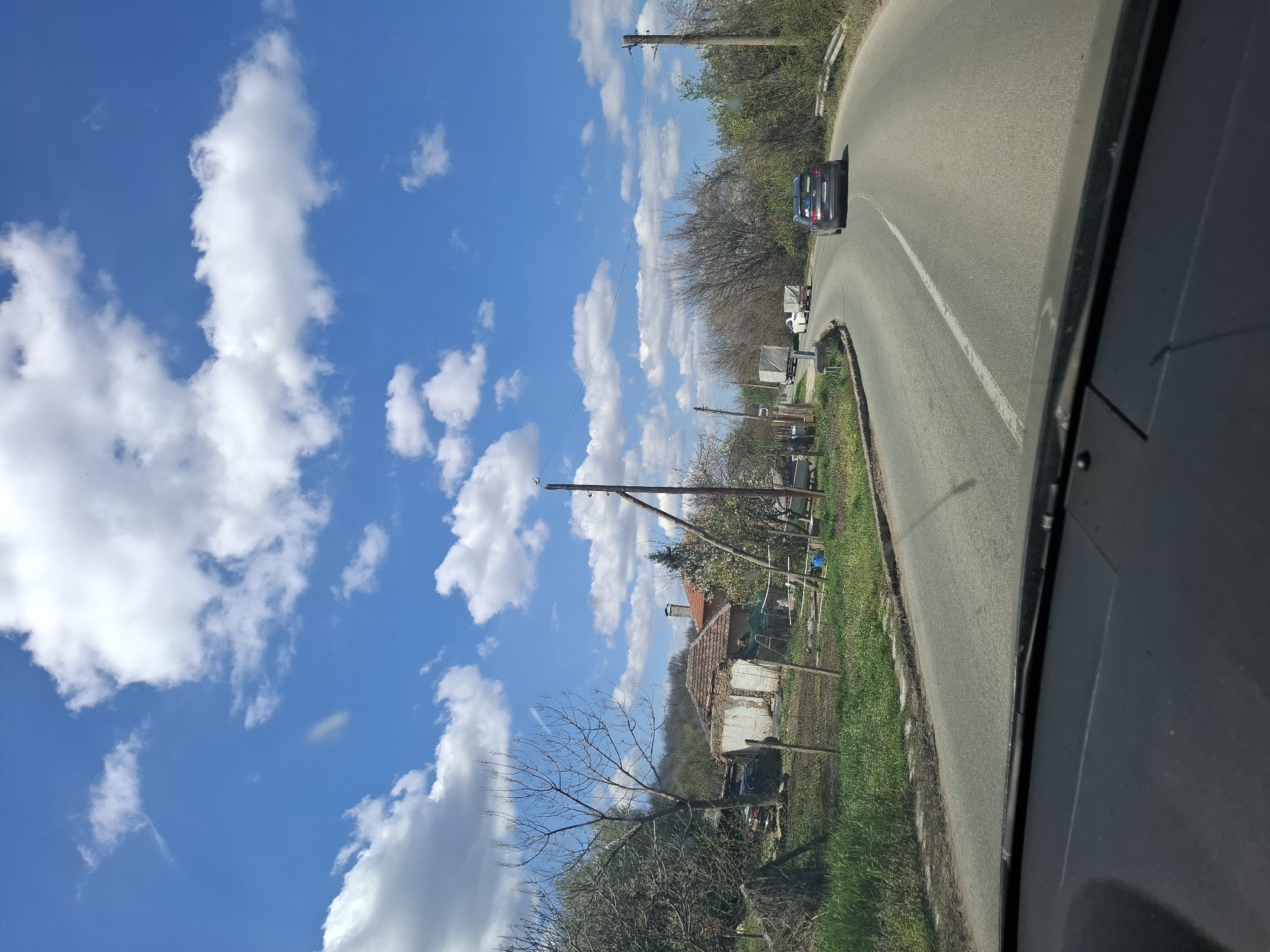 View from inside a vehicle, looking toward a winding road lined with greenery and utility poles under a blue sky with scattered clouds. A car is visible on the road ahead. On the left side of the road, there are houses and trees. The location is suggested to be near or in Tsar Simeonovo, Bulgaria.