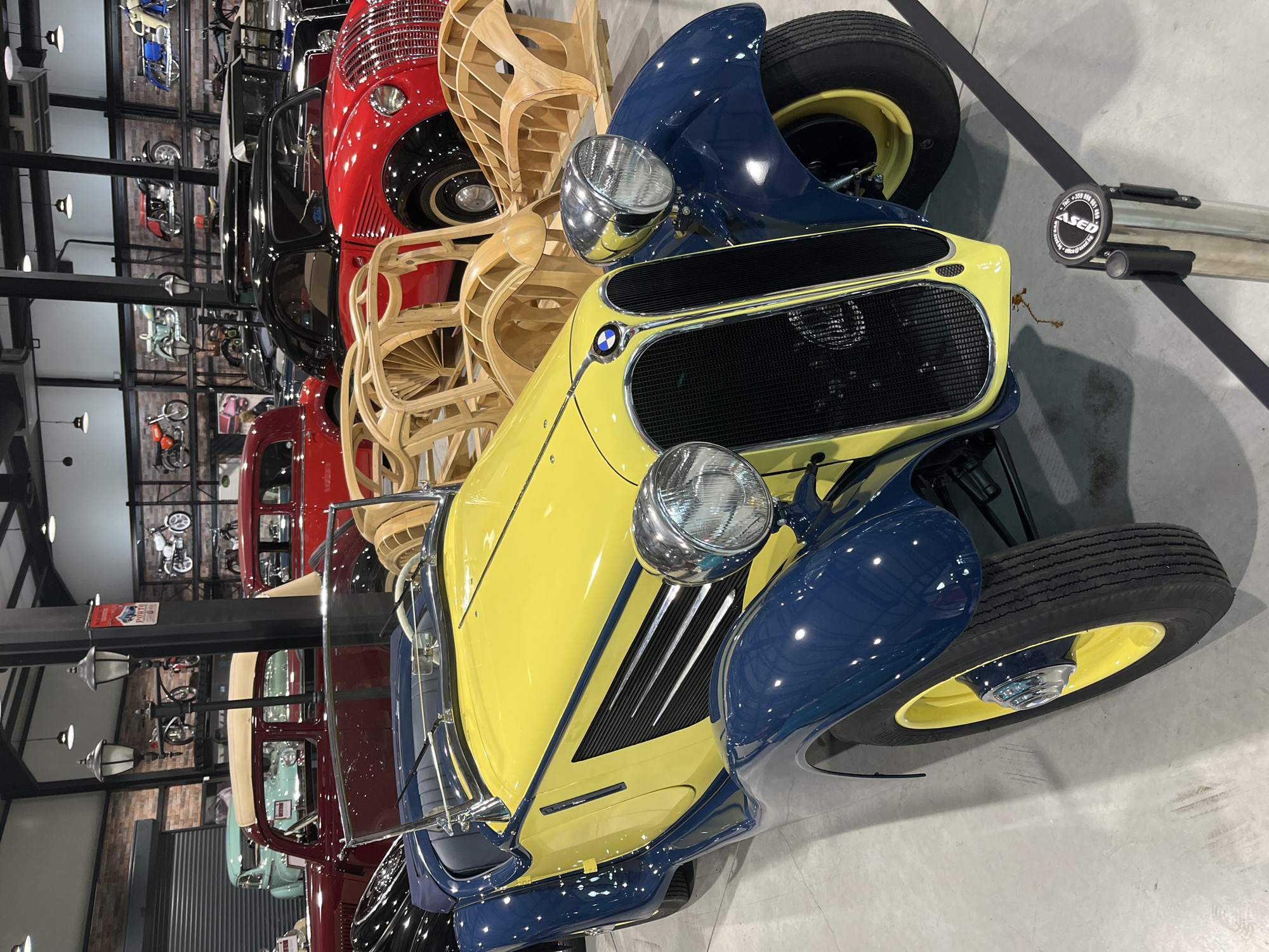 The image depicts a yellow and blue vintage BMW car in an indoor setting, possibly a museum or automotive showroom. The car is positioned at an angle, showcasing its front design with prominent headlights and a distinctive grill bearing the BMW logo. Behind the car, several other vintage vehicles are visible, indicating a collection or display of classic automobiles. A wooden, chair-like art installation is situated next to the BMW. The location appears to be a professionally lit space with a smooth, light-colored floor. Wall decor suggests a thematic automotive history display, including photographs of motorcycles.
