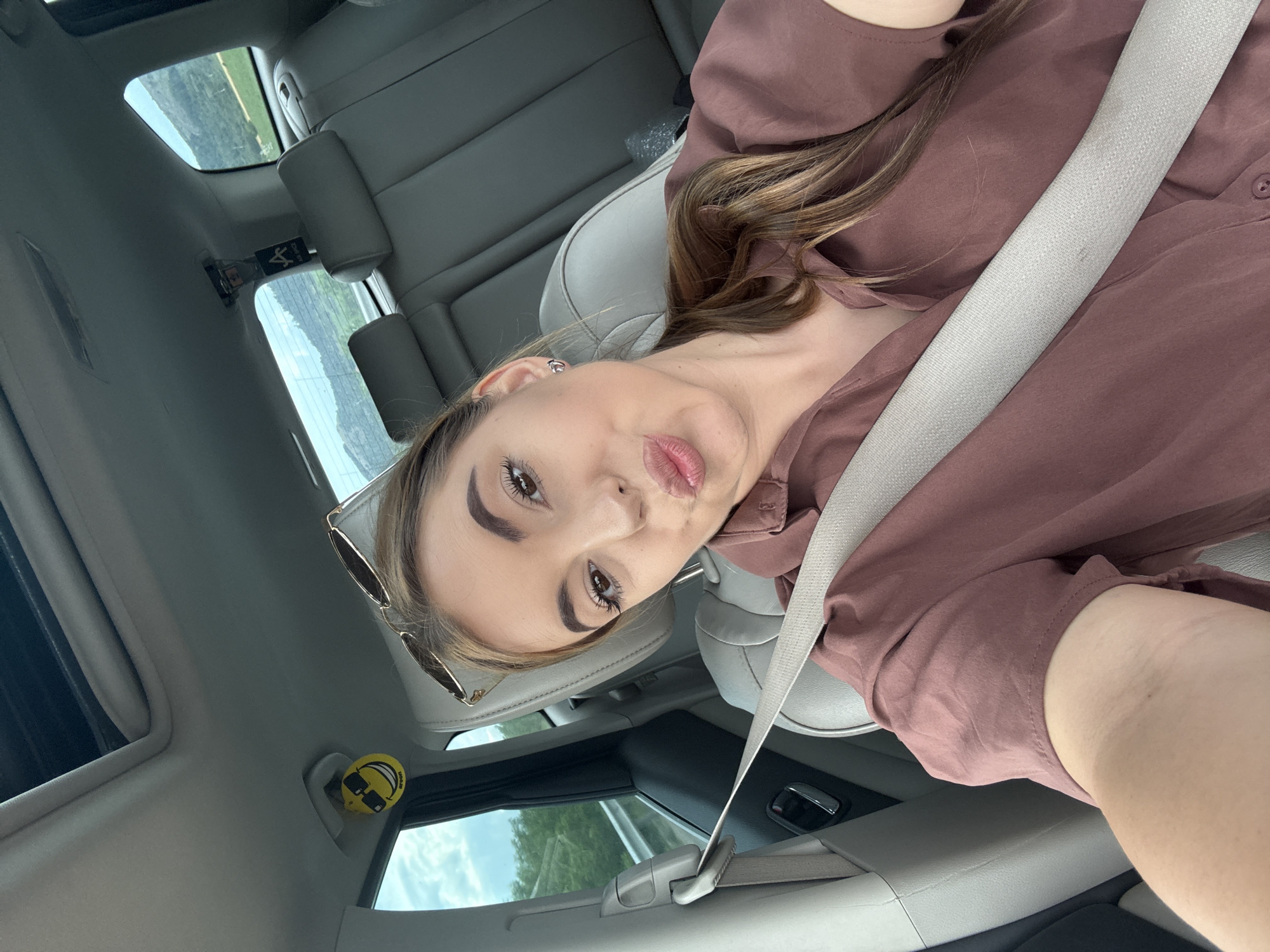 A young woman with brown hair is taking a selfie in the driver's seat of a car. She is wearing a mauve-colored button-up shirt and sunglasses are perched on her head. A seatbelt is diagonally across her body. The interior of the car is visible, including the grey leather seats, sunroof, and windows which look out onto a landscape of hills and greenery. A yellow smiley face air freshener is hanging from the rearview mirror. The car appears to be moving, as indicated by the blurred scenery outside the window. This image was taken in Aldomirovtsi, Bulgaria.
