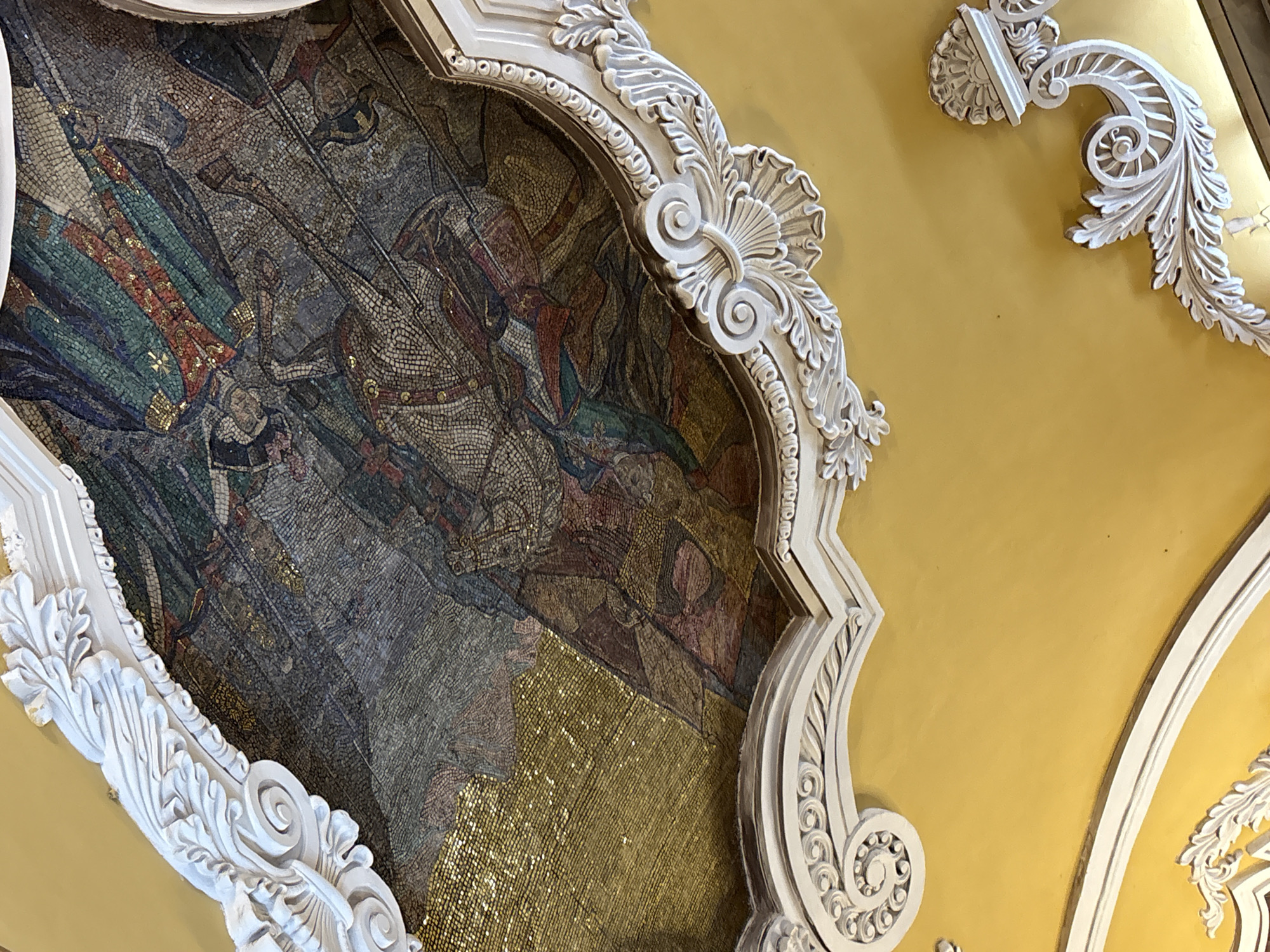 The image shows a mosaic panel framed by ornate, white plasterwork, set against a yellow wall. The mosaic depicts a historical scene with figures in armor and robes, possibly a battle or ceremonial event. The setting appears to be an interior space in a building in Moscow, Russia. The mosaic is comprised of small tesserae of various colors, including greens, browns, reds, and gold. The plasterwork frame is highly detailed with floral and scroll motifs. The yellow wall is a simple, unadorned surface. The perspective of the photograph is looking upwards towards the ceiling.