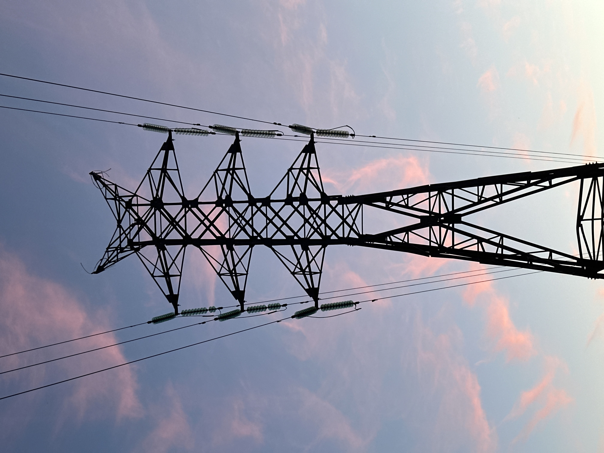 A silhouette of a high-voltage electricity pylon stands against a twilight sky. The pylon is made of dark metal lattice work, with wires extending from insulators along its structure. Wispy clouds are visible in the sky, tinged with pink and purple hues. The image is taken from a low angle, looking up at the pylon. The scene is quiet and still, evoking a sense of industrial presence within a natural setting. The specific location in the city of Perm, Russia, is not identifiable from the visual information provided.