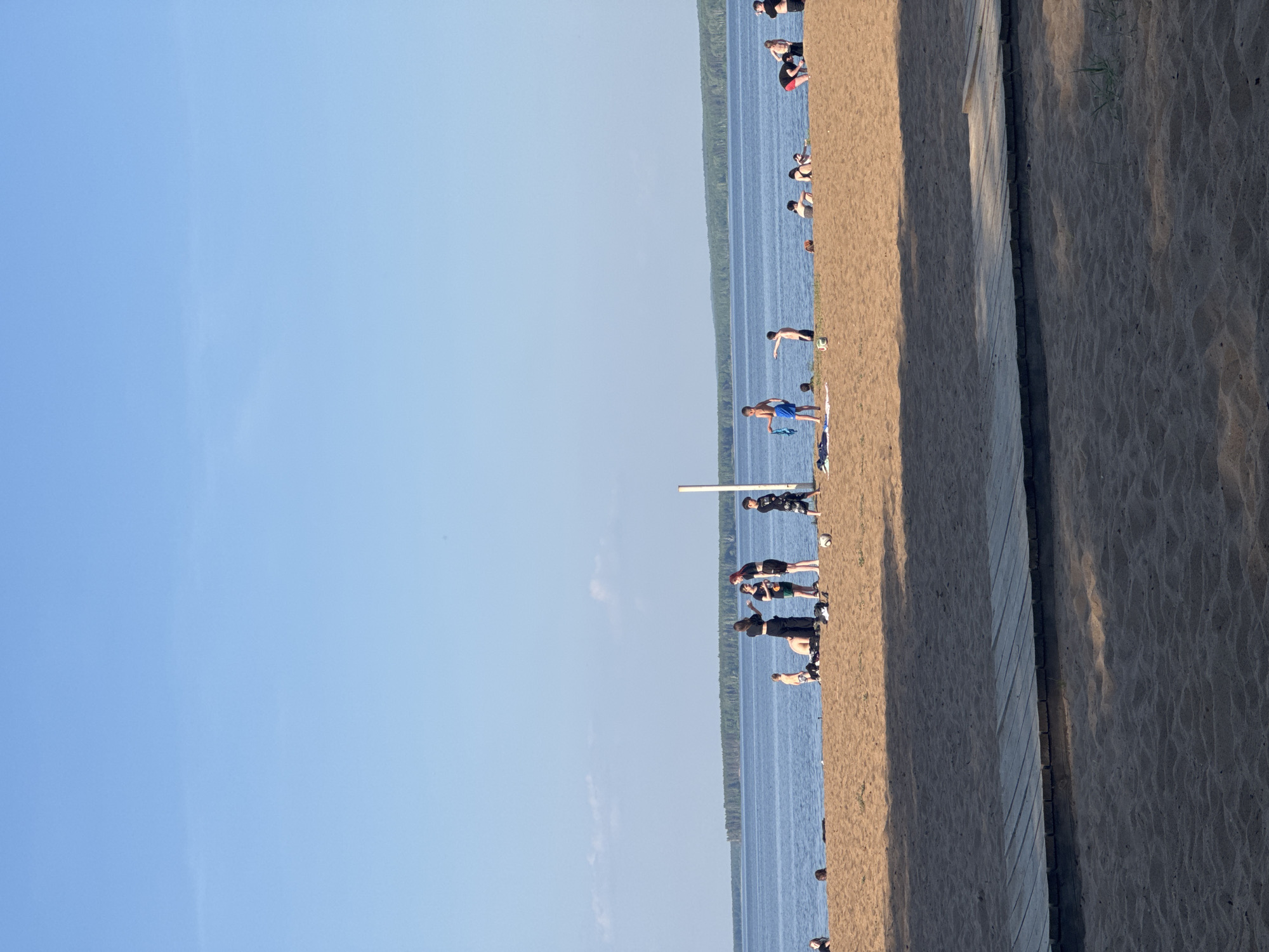The image depicts a beach scene in Chaikovsky, Russia. A group of people is scattered along the shoreline of a body of water, possibly the Kama River or a reservoir near the city. Individuals are seen standing, wading, and generally enjoying the beach. To the right of the beach is a wooden walkway or path. The sky is clear and blue.