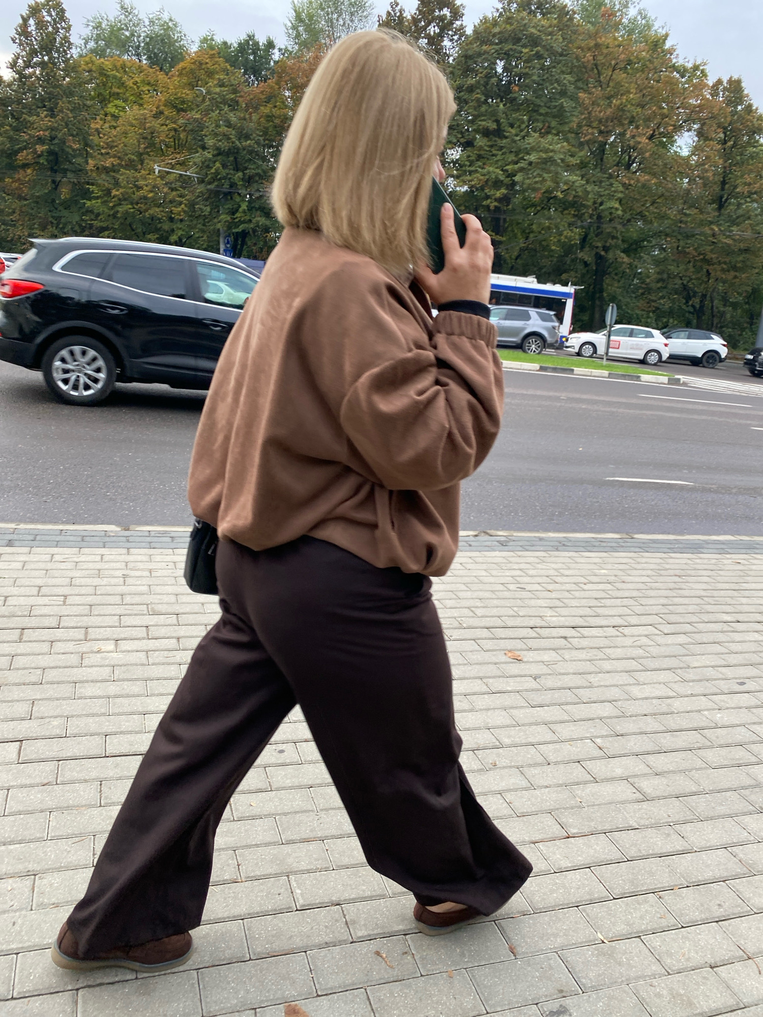 A woman with blonde shoulder-length hair is walking on a paved sidewalk and talking on a green cell phone. She is wearing a loose-fitting, brown bomber-style jacket over dark brown wide-leg pants and brown slip-on shoes. A small black purse is slung over her right shoulder. The background shows a street with several cars, including a black SUV, a silver SUV, two white cars, and a bus. Trees with green and autumn-colored foliage are visible behind the street. The sky is overcast. This scene is likely from Chișinău, Moldova, given the context.