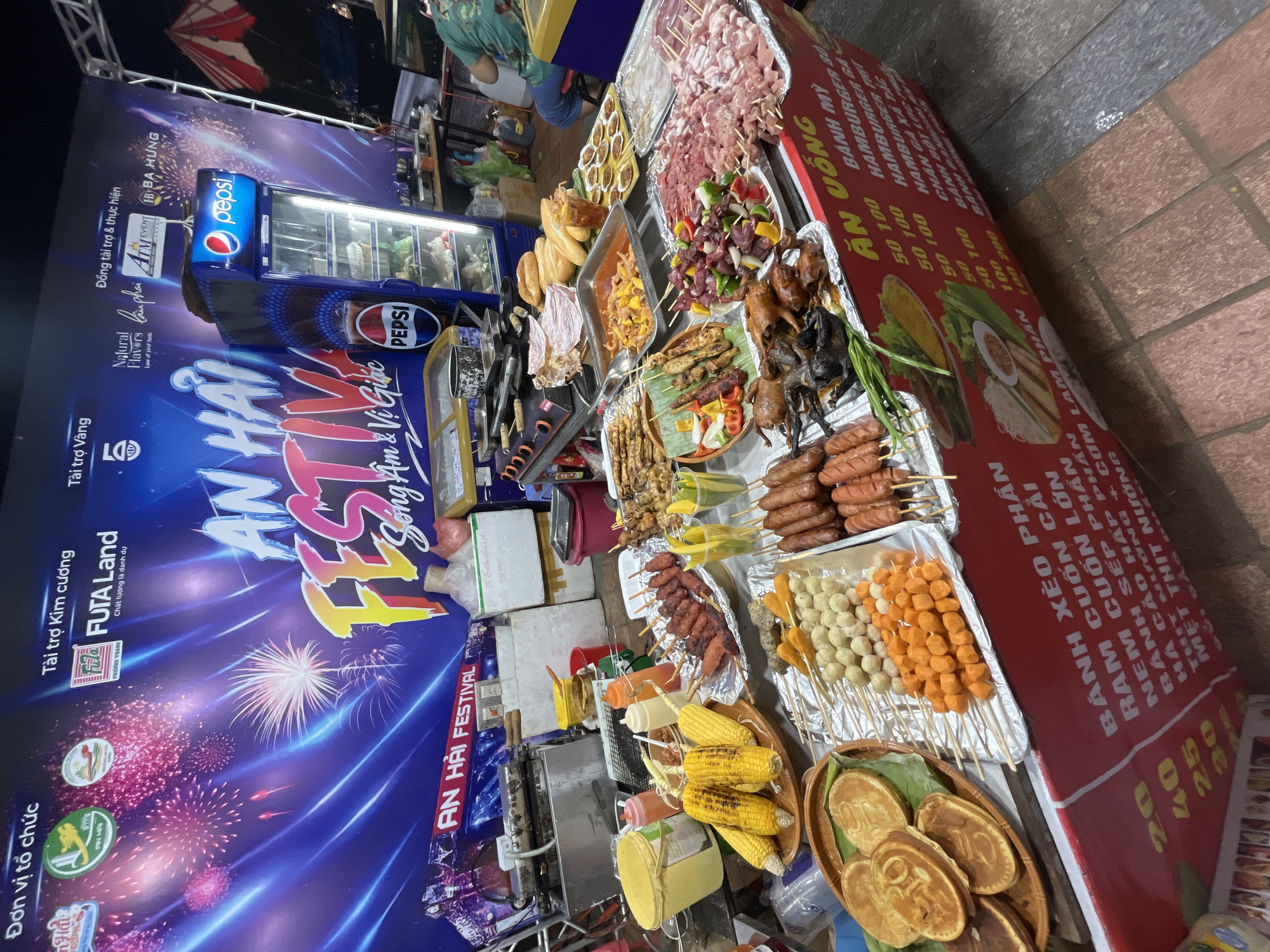 The image depicts a bustling street food stall in Da Nang, Vietnam, likely during an outdoor festival. A large banner with fireworks and the text "AN HAI FESTIVAL SONG A VI GIAC" serves as the backdrop, indicating a festive event. In the foreground, a red food stall displays an array of grilled meats, seafood, sausages, and corn on the cob, all arranged on skewers and platters. Several types of skewers are visible, including chicken, pork, and possibly seafood meatballs. Various sauces and condiments are also present. Behind the stall, a Pepsi refrigerator is stocked with drinks. A person in a green shirt is partially visible in the background, seated at another stall. The ground is paved with reddish-brown bricks. The overall scene suggests a lively and food-centric atmosphere typical of Vietnamese street markets or festivals.