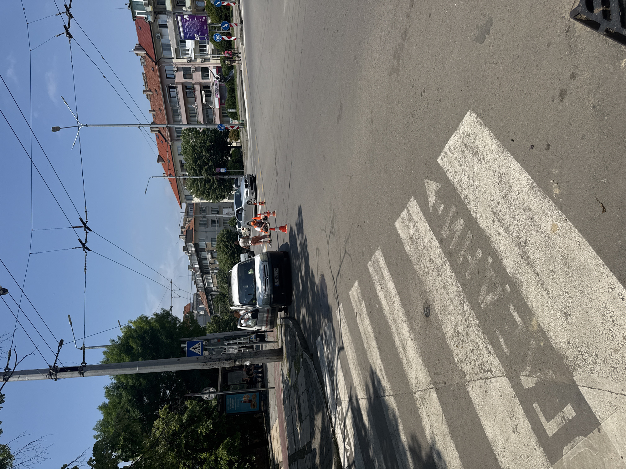 The image captures an urban street scene on a clear, sunny day, likely mid-morning or afternoon, indicated by the bright light and long shadows cast by objects and buildings. The perspective is angled downwards towards a road intersection.
In the foreground, a distinct pedestrian crossing with white zebra stripes is visible on the asphalt. Shadows from nearby trees or structures fall across these markings. To the right, part of a black metal drain grate can be seen.
The central focus is a white van, possibly a Citroën Berlingo, parked near the curb. Its license plate, "B 4078 PM", is visible, suggesting a location in Sofia, Bulgaria. Two individuals are near the van; one is wearing orange high-visibility work attire and appears to be involved in some activity, possibly road maintenance or utility work, given the presence of orange traffic cones around the vehicle and a ladder leaning against the curb or the van. Another person is also near the van. Further down the road, a white sedan, possibly a BMW, is also parked.
The background features multi-story buildings with European-style architecture, some with tiled roofs. Overhead, numerous power lines or trolleybus cables crisscross the clear blue sky. Street light poles are also visible, along with lush green trees. Various road signs are scattered throughout the scene, including blue circular signs indicating mandatory directions (e.g., turn right, straight ahead) and a blue square sign denoting a pedestrian crossing. A red circular sign with a diagonal line (e.g., no entry or no parking) is also present. On a lamppost near the sidewalk, a blue advertising poster with some text and an image of food (possibly a burger) can be partially seen. Road markings with obscured text, possibly "STOP," are visible on the street within the pedestrian crossing.