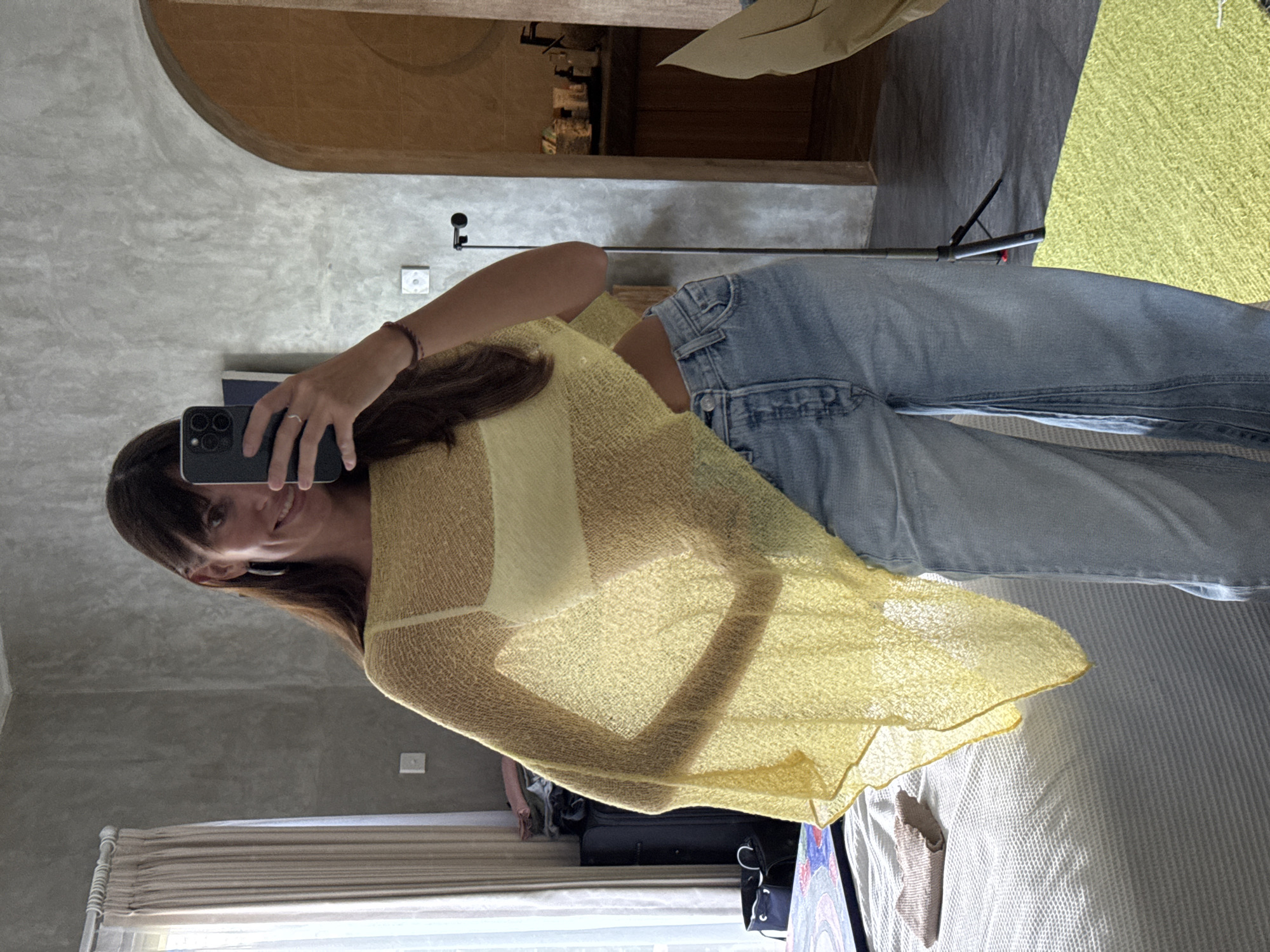 The image is a mirror selfie, oriented sideways (rotated 90 degrees counter-clockwise). It depicts a young woman with long, dark brown hair and bangs, captured from the chest down, reflecting in a large mirror. She is holding a dark-colored smartphone with triple cameras in her left hand, taking the photo, and displays a faint smile. She is wearing a light yellow, sheer, knitted top or cardigan over a lighter undergarment, paired with light blue denim jeans. A dark-colored bracelet is visible on her right wrist, and a ring on her left ring finger.
The reflected background reveals an indoor setting, likely a modern bedroom or living area. The walls appear to be light grey concrete or stucco. An arched, dark wooden or concrete structure is visible in the upper part of the reflection. The floor is dark grey, partially covered by a bright lime green rug/mat. A bed with white and patterned bedding is situated in the lower right of the reflection. Light-colored curtains are drawn over a window. Other objects visible include a black suitcase or bag on the floor, and a black light stand or tripod partially seen behind the subject's right leg. The room is well-lit, suggesting it is daytime. The contemporary architectural style aligns with the provided location context of North Kuta, Indonesia. No visible text is present in the image.