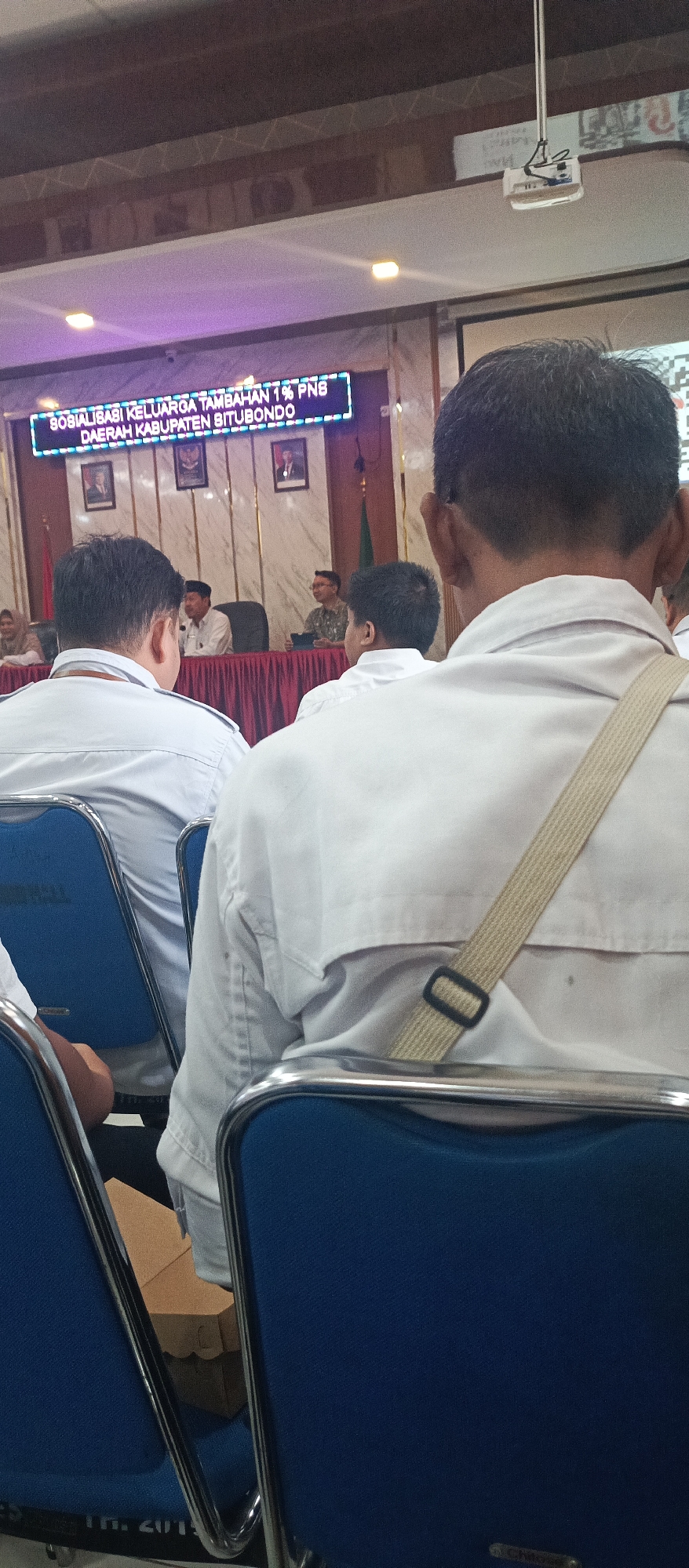 The image shows a rear view of people seated in blue chairs, facing a stage area with a screen displaying text that reads "SOSIALISASI KELUARGA TAMBAHAN 1% PNS DAERAH KABUPATEN SITUBONDO". This suggests a public meeting or seminar related to family and civil servants in the Situbondo Regency. The setting appears to be an indoor venue, possibly a conference hall or auditorium. People are wearing light-colored shirts, and some have lanyards. The lighting is a mix of ambient and artificial, with purple lights illuminating the screen. There is a projector mounted on the ceiling. The overall atmosphere is formal, indicating an official event.