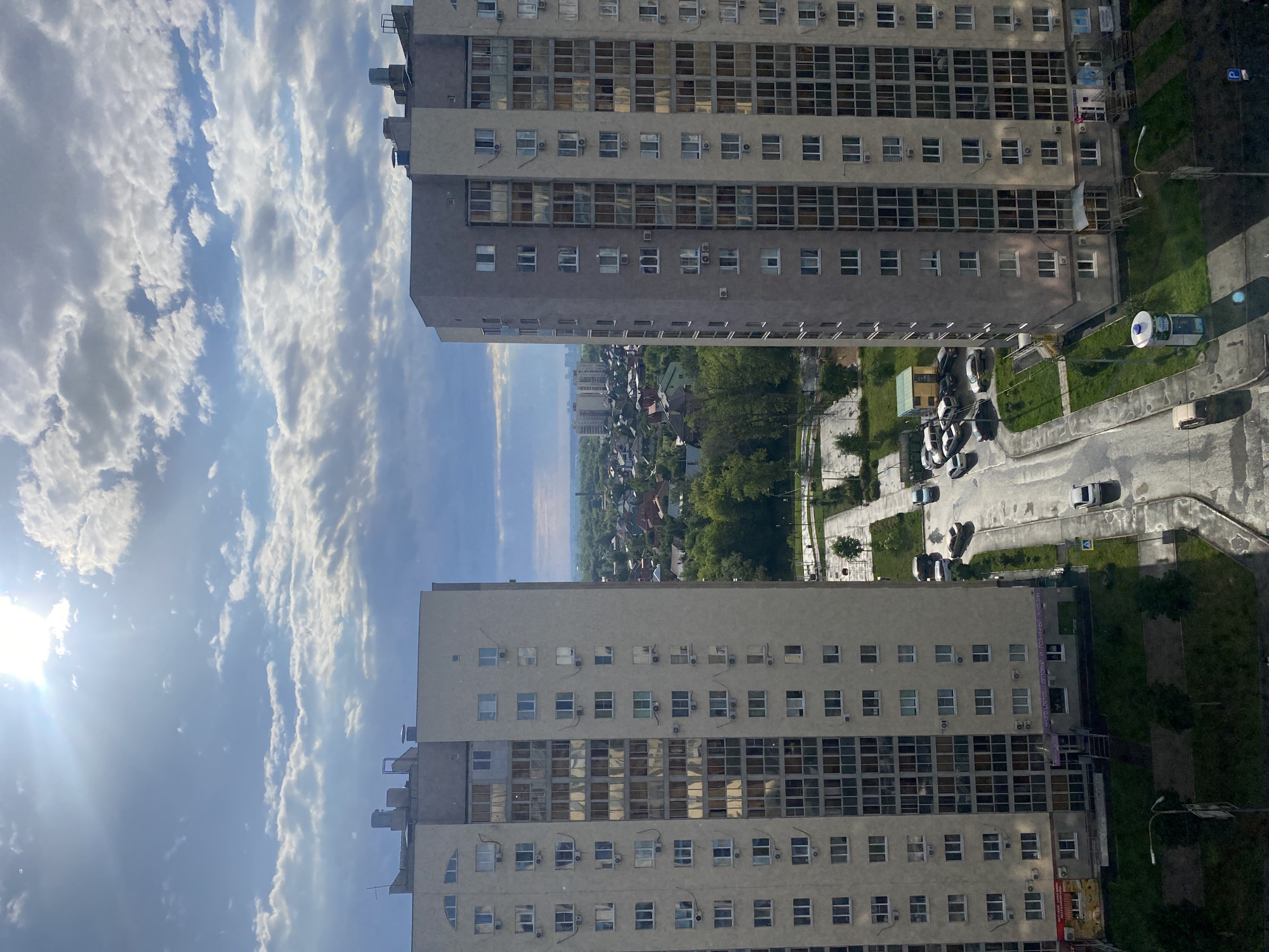 The image is an aerial view of a residential area in Novinki, Russia, on a cloudy day with the sun shining through. Two multi-story apartment buildings dominate the foreground, one on the left and one on the right, with a street and some green spaces in between. The buildings are light-colored with numerous windows and air conditioning units visible.
In the middle ground, there is a street with several cars parked and driving. Beyond the street, there are more residential buildings, some appearing to be houses with red roofs, interspersed with trees. The background shows a hazy cityscape with more apartment buildings and a distant horizon. The sky is partly cloudy, with the sun creating lens flare in the upper left portion of the image. The overall impression is of a somewhat dense urban environment with pockets of greenery. There are no people clearly visible in the image.