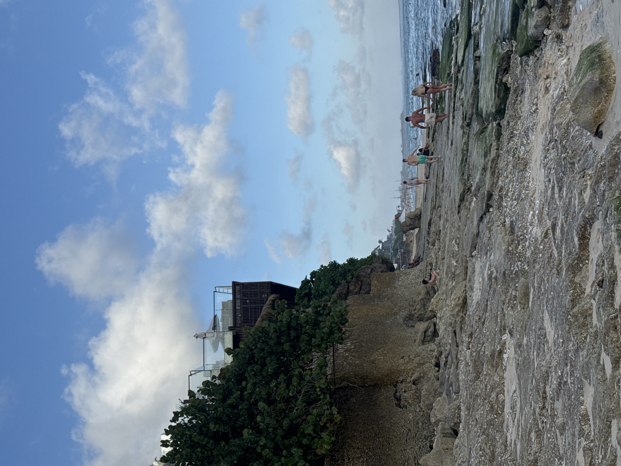 The image depicts a coastal scene, likely in Kuta Selatan, Indonesia, based on the environment and architecture. The sky is a mix of blue and white clouds, with a building complex visible on the left, partially obscured by dense green foliage. The structure appears to be tiered, with possible balconies.
A rocky shoreline dominates the right side of the image, leading down to the ocean. Several individuals are present on the rocks, some standing near the water's edge. These individuals appear to be dressed in swimwear or casual attire. A larger rock is covered with vegetation.
The ocean is in the background. The focus of the image is on the rocky landscape, the people exploring it, and the building perched above.