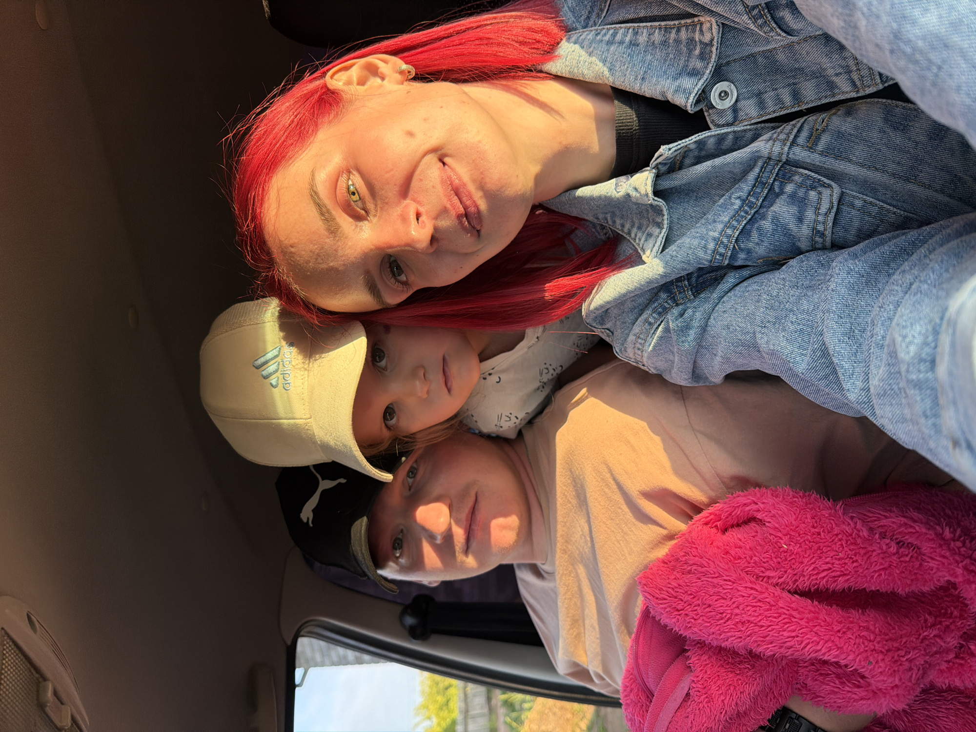 A rotated, brightly lit selfie captures three individuals inside a car, likely in Chaikovsky, Russia, during golden hour. Strong, warm sunlight illuminates their faces from the right, suggesting late afternoon or early morning.
At the top of the frame, a woman with vibrant red hair and striking green/hazel eyes smiles gently at the camera. She wears a light blue denim jacket over a black top, with an earring visible in her left ear. Beneath her, a child with light brown hair looks directly at the camera with a neutral expression. The child wears a light beige baseball cap with an "Adidas" logo and a white top featuring subtle patterns. Below the child, another individual with light brown hair and a neutral gaze wears a light pink or peach t-shirt and a dark baseball cap, where a "Puma" logo is visible. A bright pink fuzzy jacket or blanket is visible on their lap or beside them.
The setting is inside a car, with parts of the ceiling and a car window showing a sunny outdoor scene visible in the background. The subjects appear to be posing for a casual group photo within the vehicle.