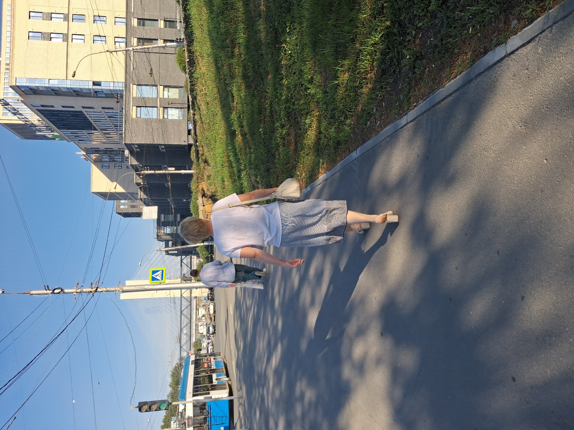 Two people, a woman and a man, are walking away from the camera on a paved pedestrian path adjacent to a street in Novosibirsk, Russia. The woman, in the foreground, wears a white t-shirt and a striped skirt, carrying a light-colored purse. The man, behind her, wears a light grey long-sleeved shirt and dark pants, carrying a white shopping bag. A blue and white trolleybus is visible on the street to the left, and several cars are proceeding in the distance. Modern, multi-story buildings, some with brick facades and large windows, line the opposite side of the street. A suspension bridge and a tall traffic signal pole with overhead wires are also present in the background. The scene is illuminated by clear daylight, casting long shadows across the path.