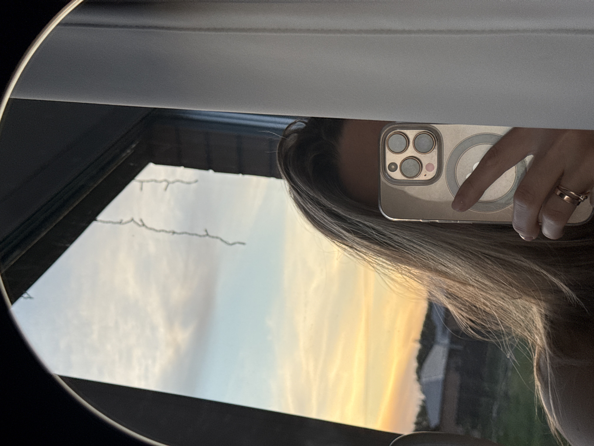 A person is taking a selfie in a round mirror. The reflection in the mirror shows a sunset sky, a building, part of the person's face, and their hand holding an iPhone. The location could not be determined.