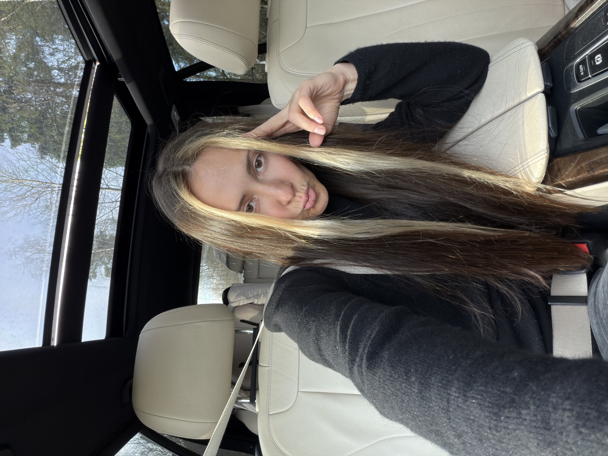 A woman is in the passenger seat of a car. She is wearing a black sweater and a seatbelt, with long brown hair with blonde streaks and she is making a hand gesture near her forehead. The car has a sunroof and beige leather interior. The location appears to be Babeevo, Russia, judging by the trees visible through the windows.
