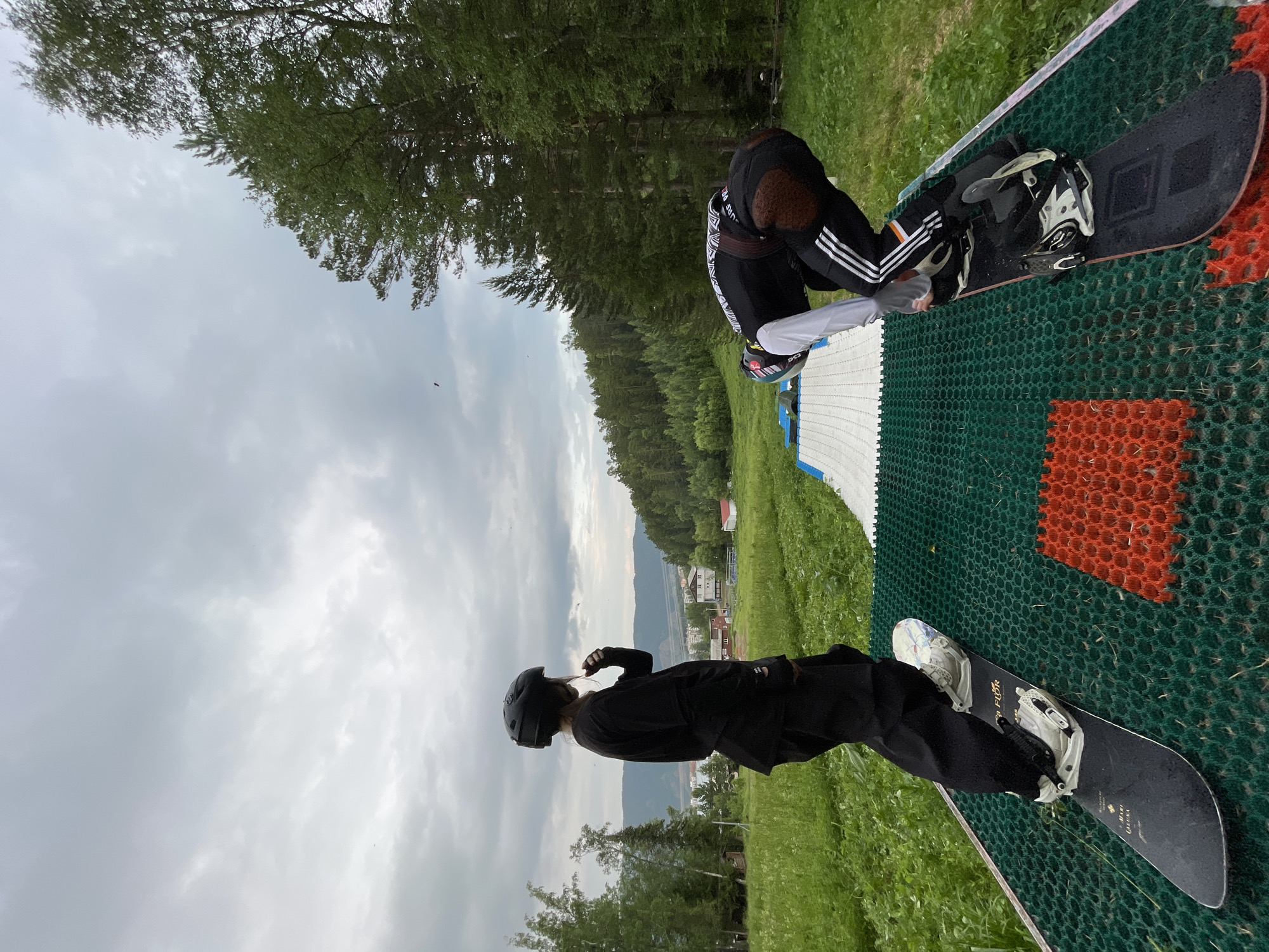 Two individuals wearing snowboarding gear are positioned on a green mat with a textured surface, each with a snowboard attached to their feet. The person on the left stands upright, facing away from the camera and looking towards a distant landscape. The person on the right is in a crouched position, preparing to snowboard. The mat is situated on a grassy incline, with a white ramp-like structure extending from it. In the background, there is a lush green forest, a small town with buildings, and mountains under a cloudy sky. This scene is located in Divnogorsk, Russia, a city known for its scenic beauty and outdoor recreational opportunities, including those offered by the nearby Krasnoyarskoye Reservoir.